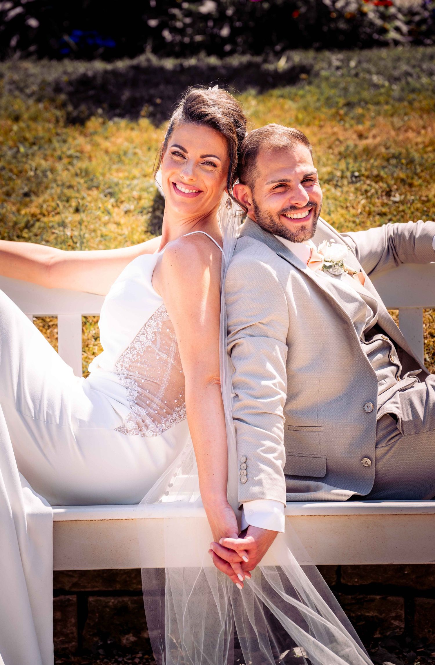 Happy bride and groom sitting back to back on white bench outdoors, holding hands and smiling.