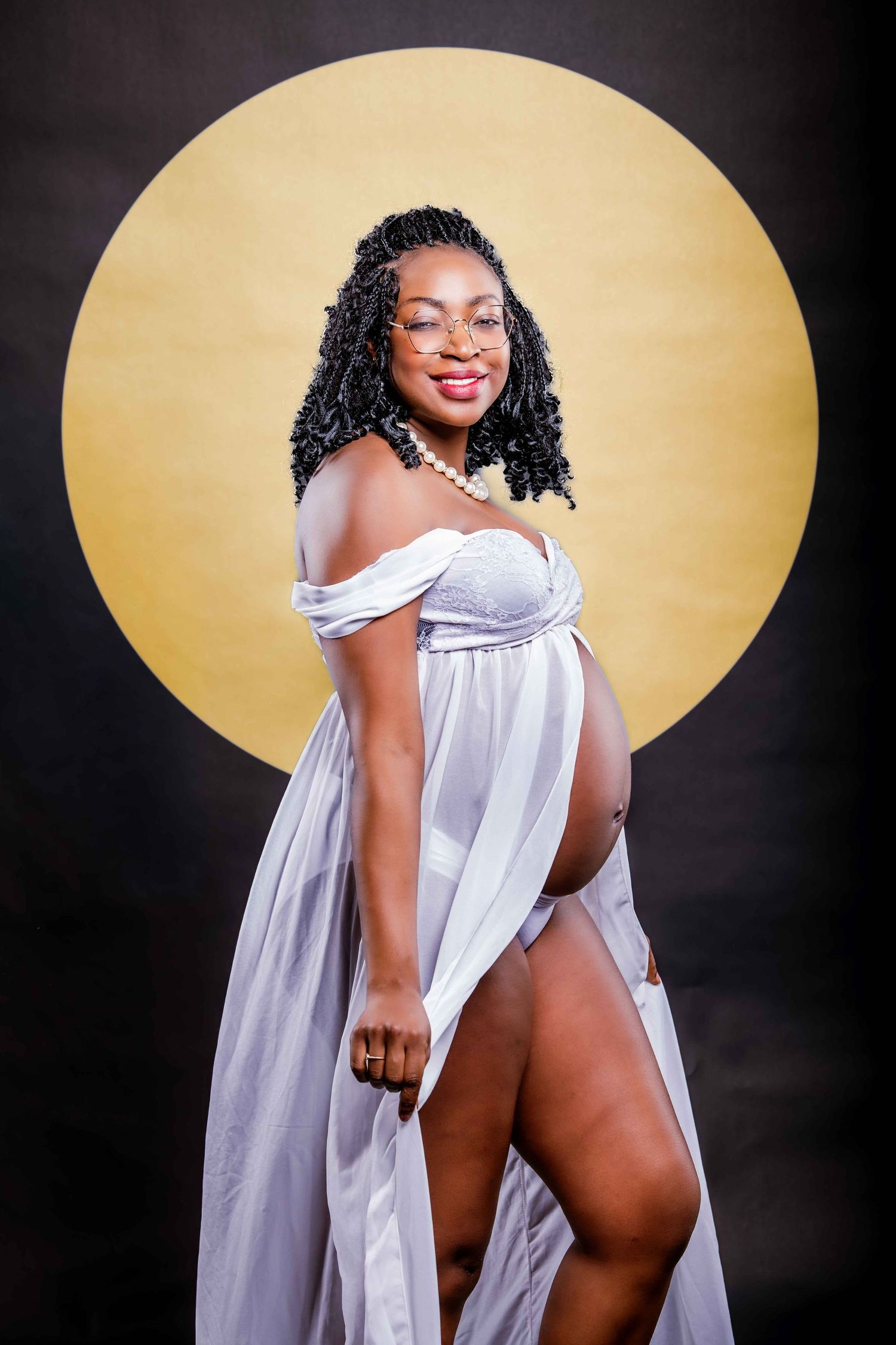 Maternity photo of an expectant mother in a flowing white off-shoulder dress posing against a golden circular backdrop.