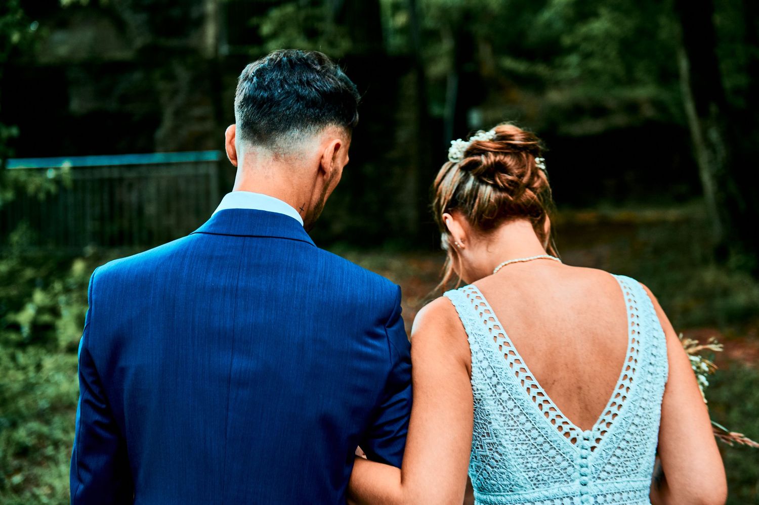 A couple in blue formal attire walking together through a lush green outdoor setting during a special event.