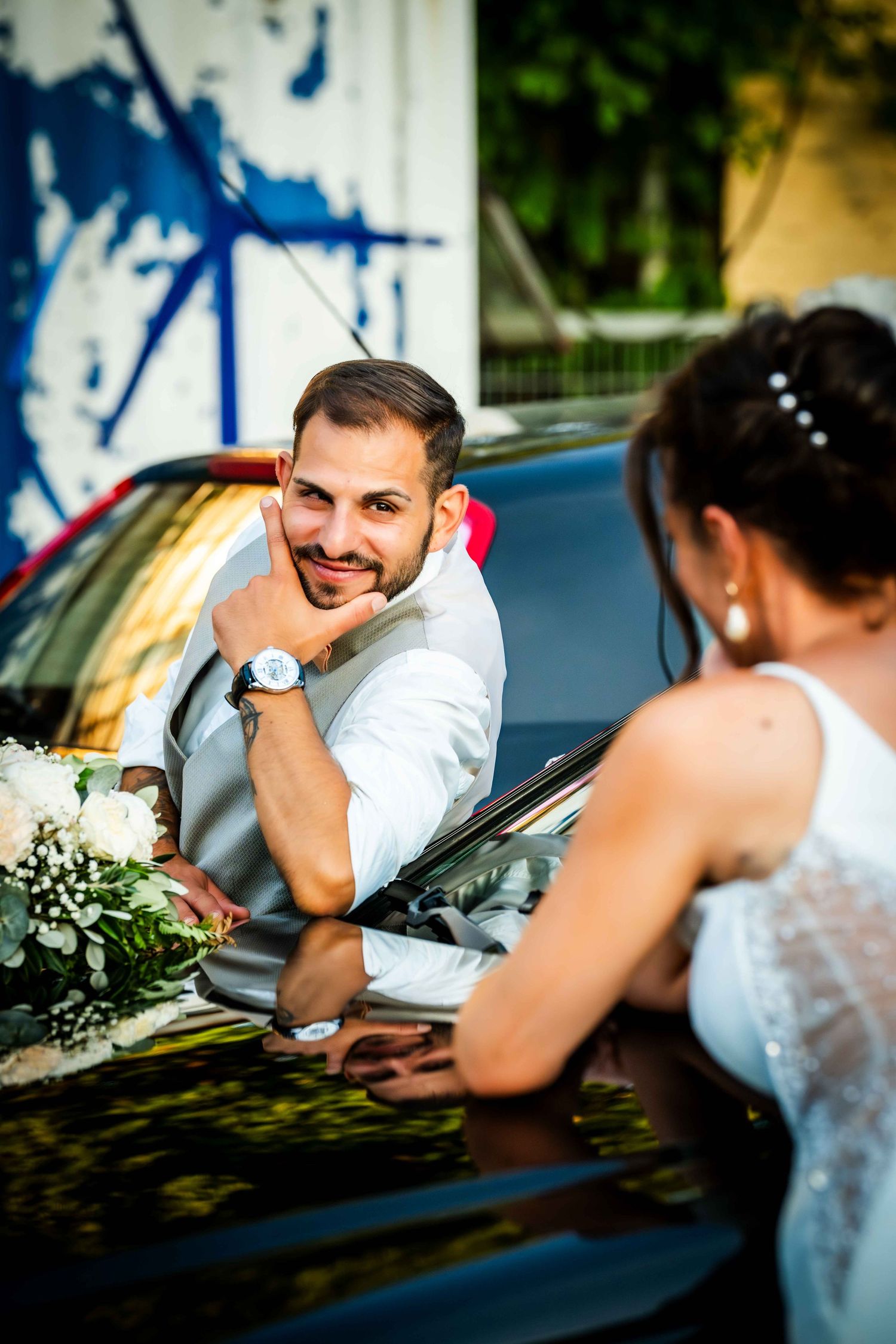 Smiling groom in gray vest leaning on black car roof looking at bride holding white wedding bouquet.