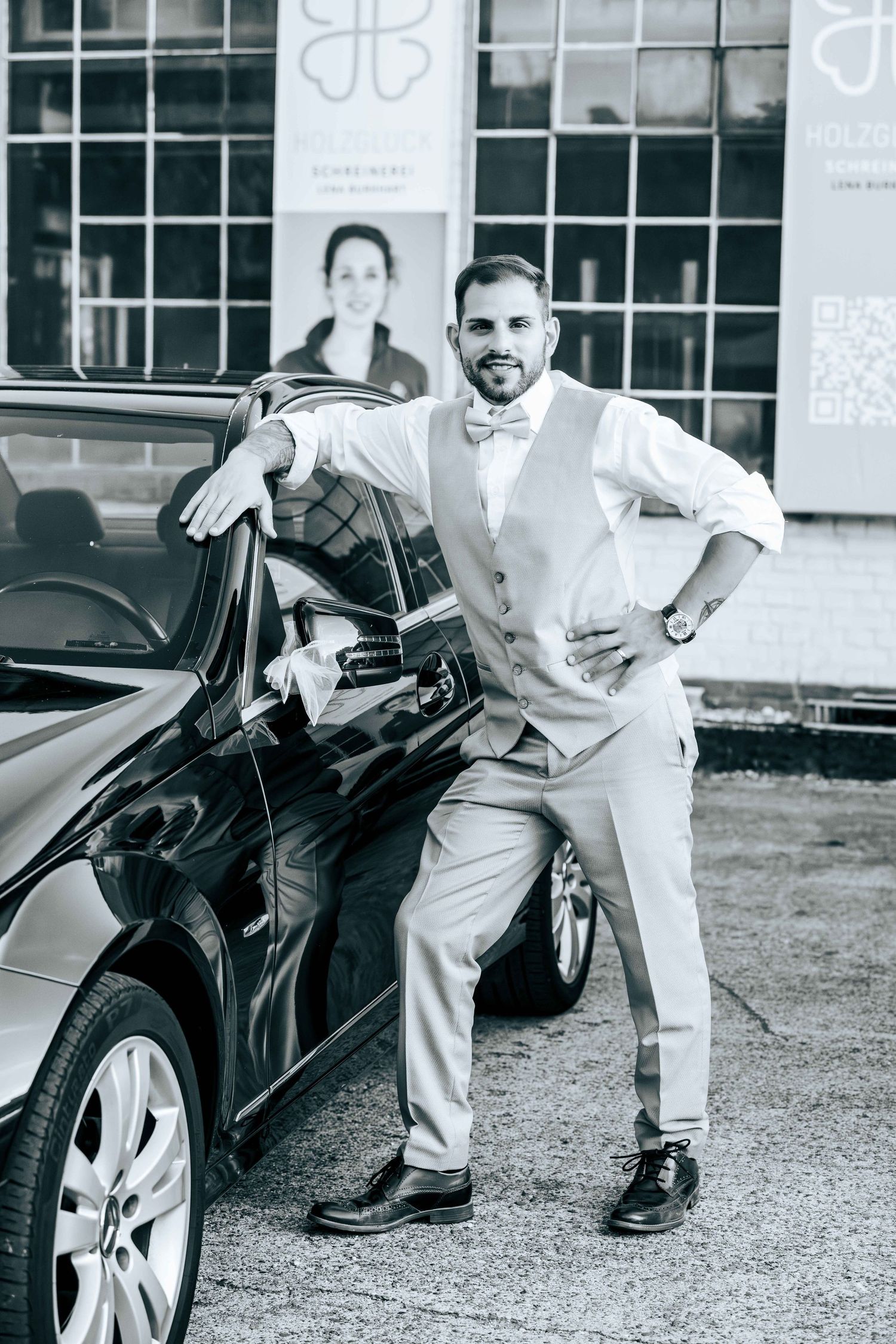Elegant groom in gray vest and bow tie leaning against a black car in a black and white wedding photo.