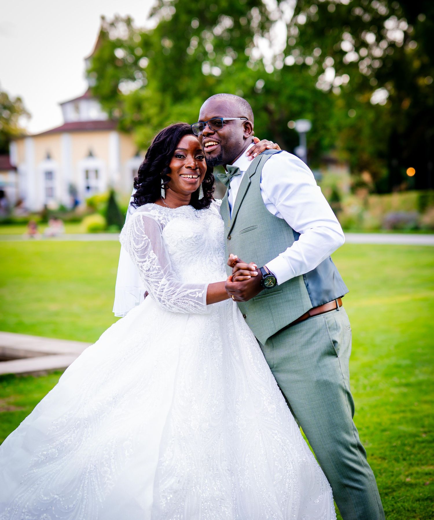 A joyful couple in wedding attire poses outdoors, the bride in a white lace gown and groom in a sage suit.
