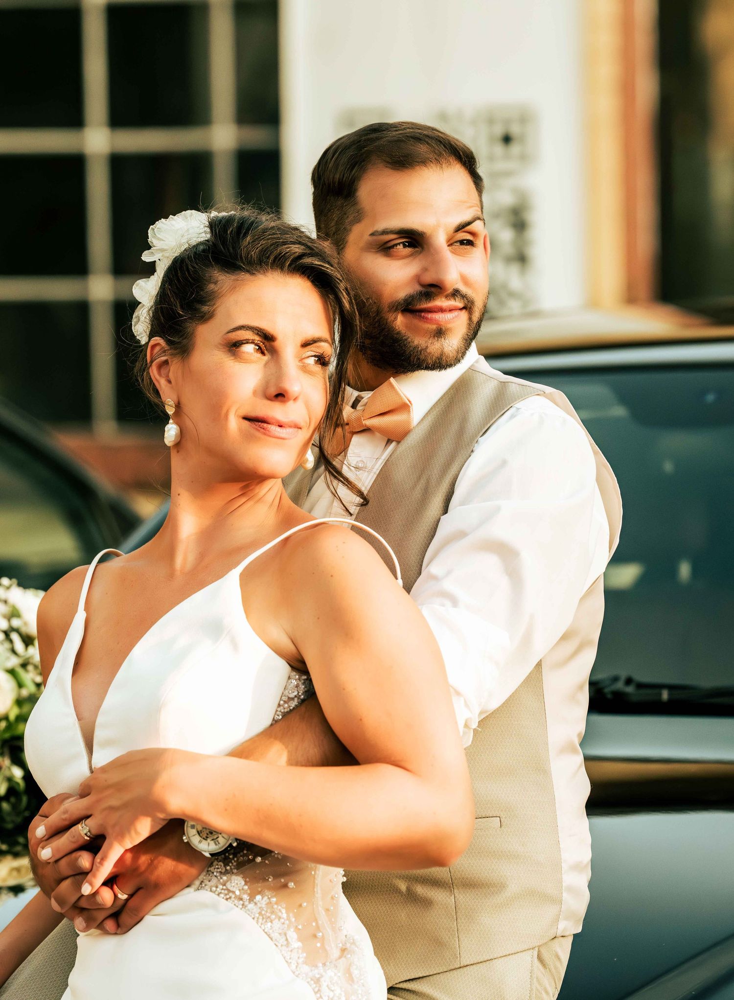 Bride and groom embrace outdoors in elegant wedding attire, groom in tan vest and bow tie, bride in white gown.