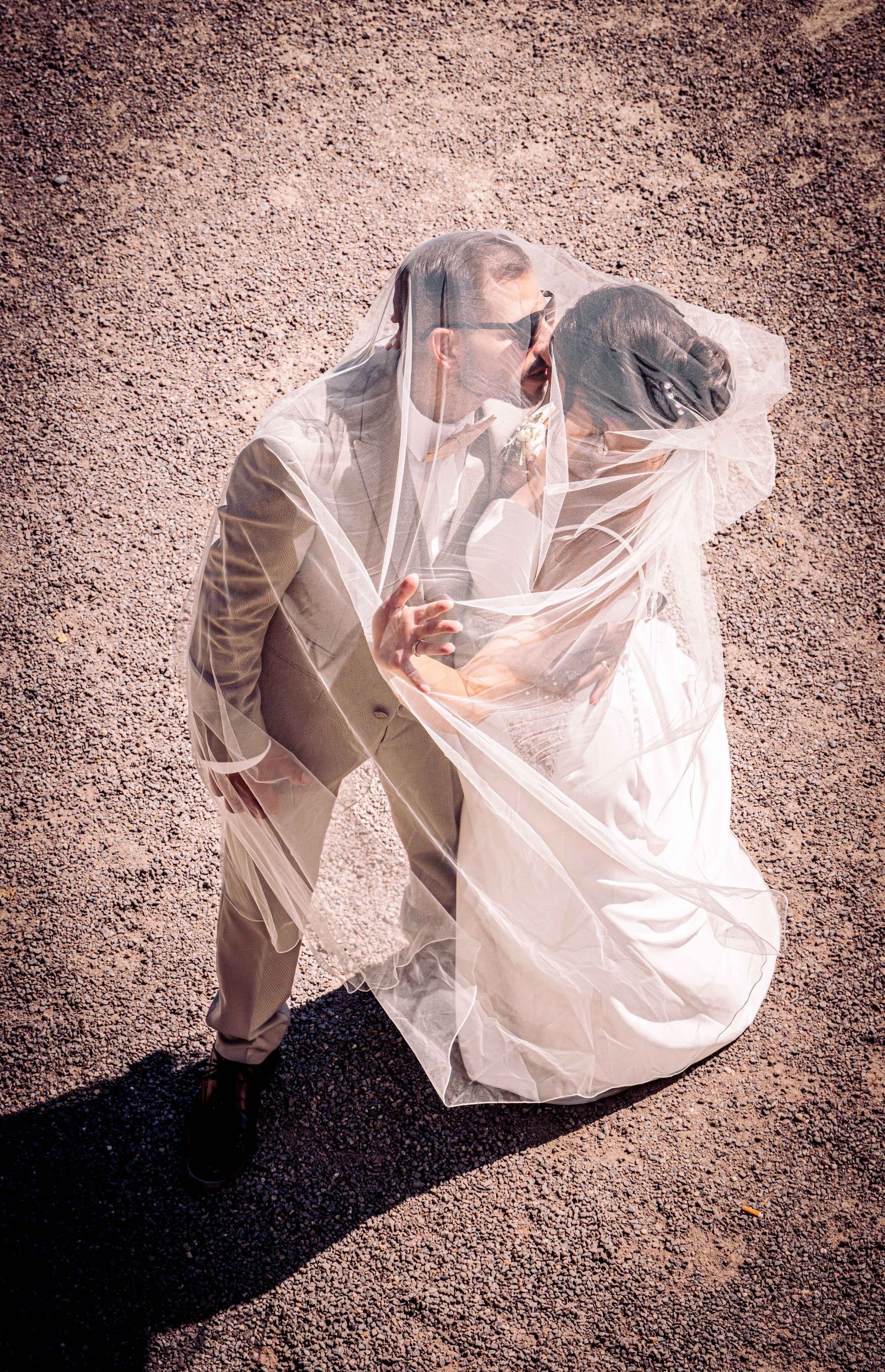 Bride and groom sharing a romantic kiss under a flowing veil, captured from above on their wedding day.