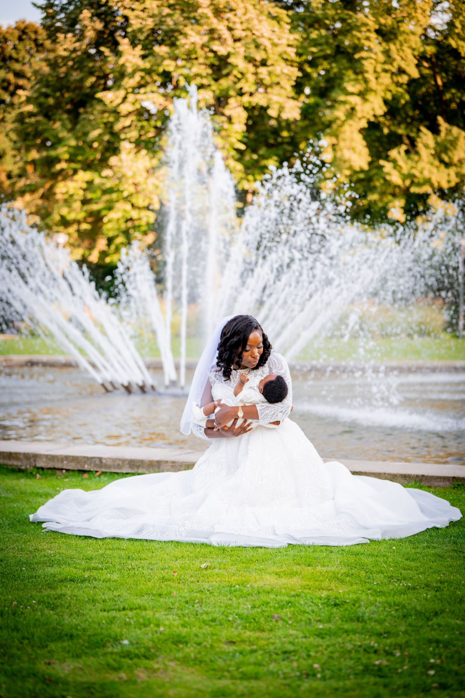 Bride in white gown sits on grass holding newborn baby in front of a fountain with autumn trees.