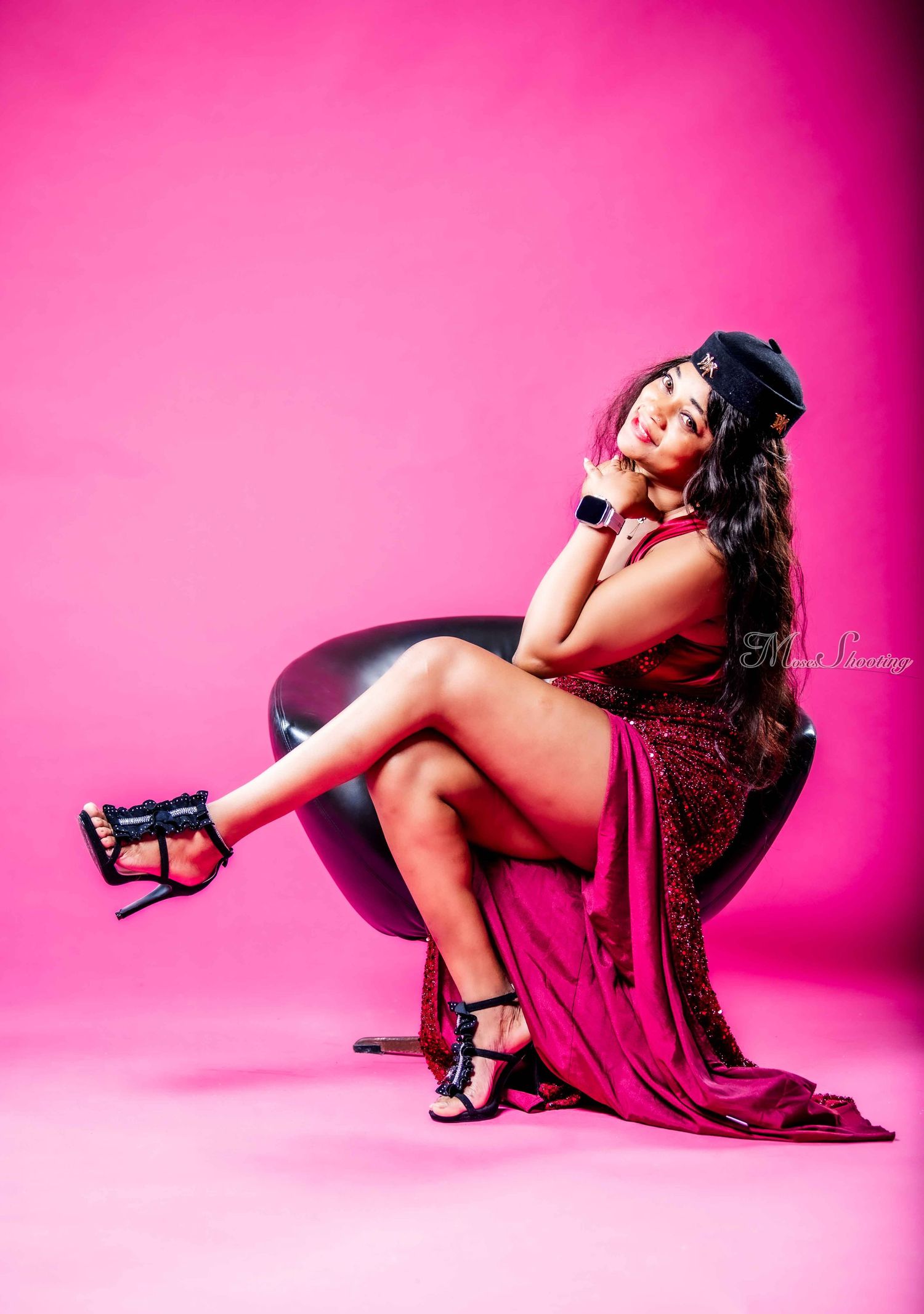 Glamorous portrait against pink backdrop features model in burgundy dress and black hat posing on modern curved chair.