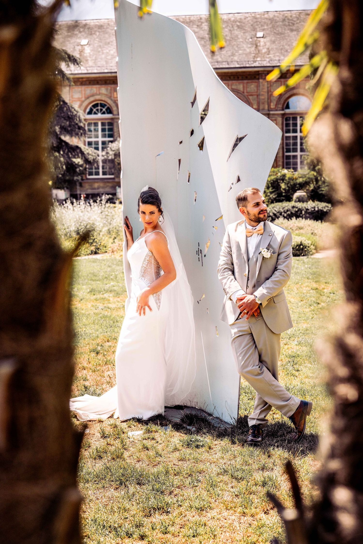 Bride and groom posing elegantly beside a white sculpture in a sunlit garden on their wedding day.