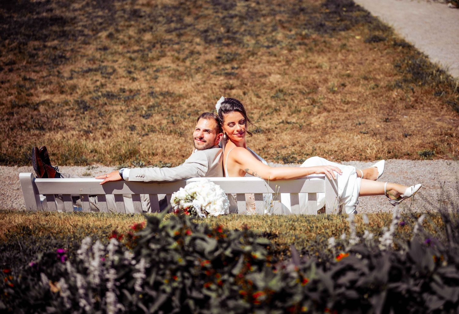 A couple relaxes together on a white outdoor bench surrounded by dry grass and flowering shrubs.