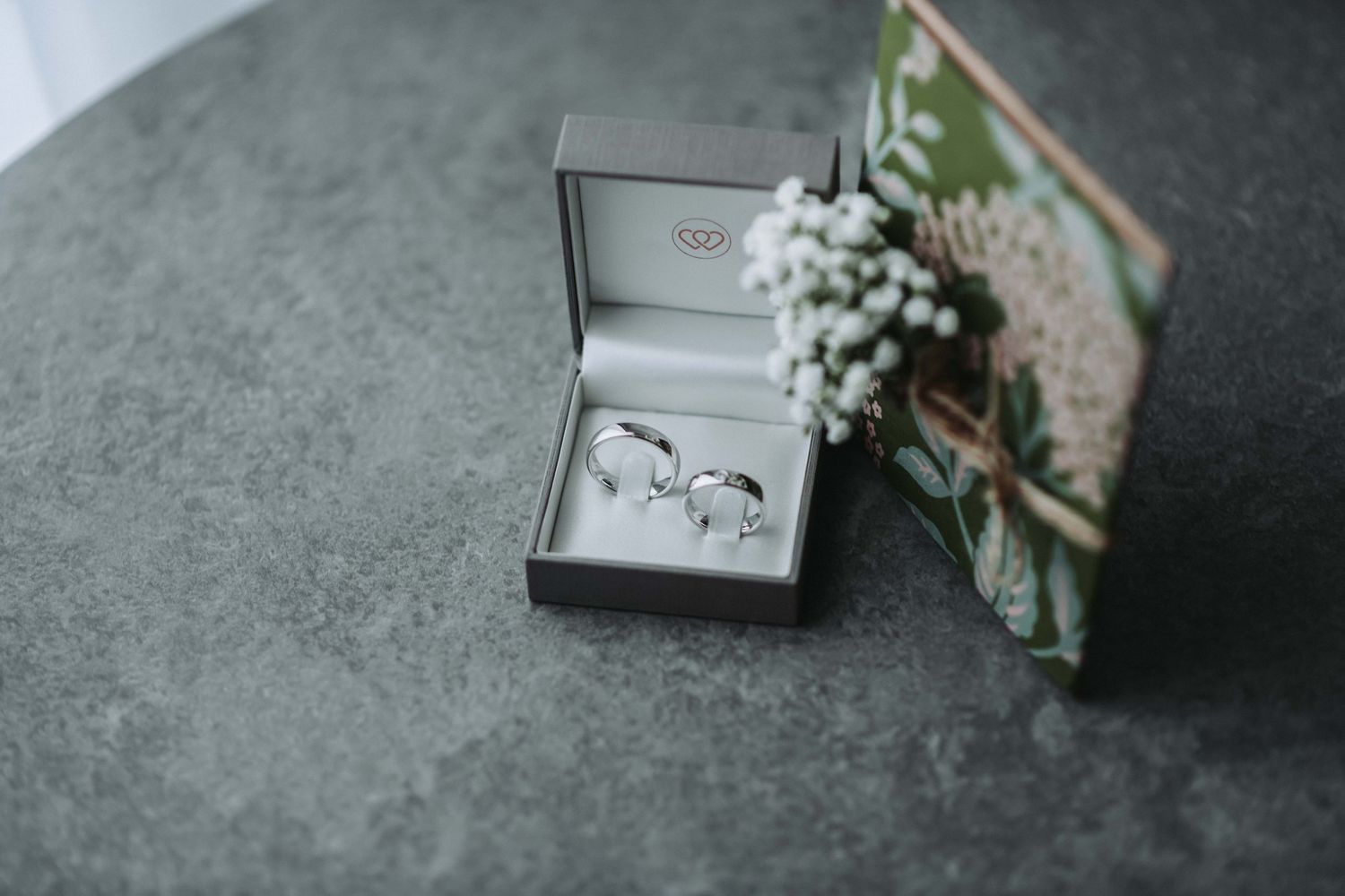Wedding rings nestled in a gray box with baby's breath flowers and a card on textured concrete surface.