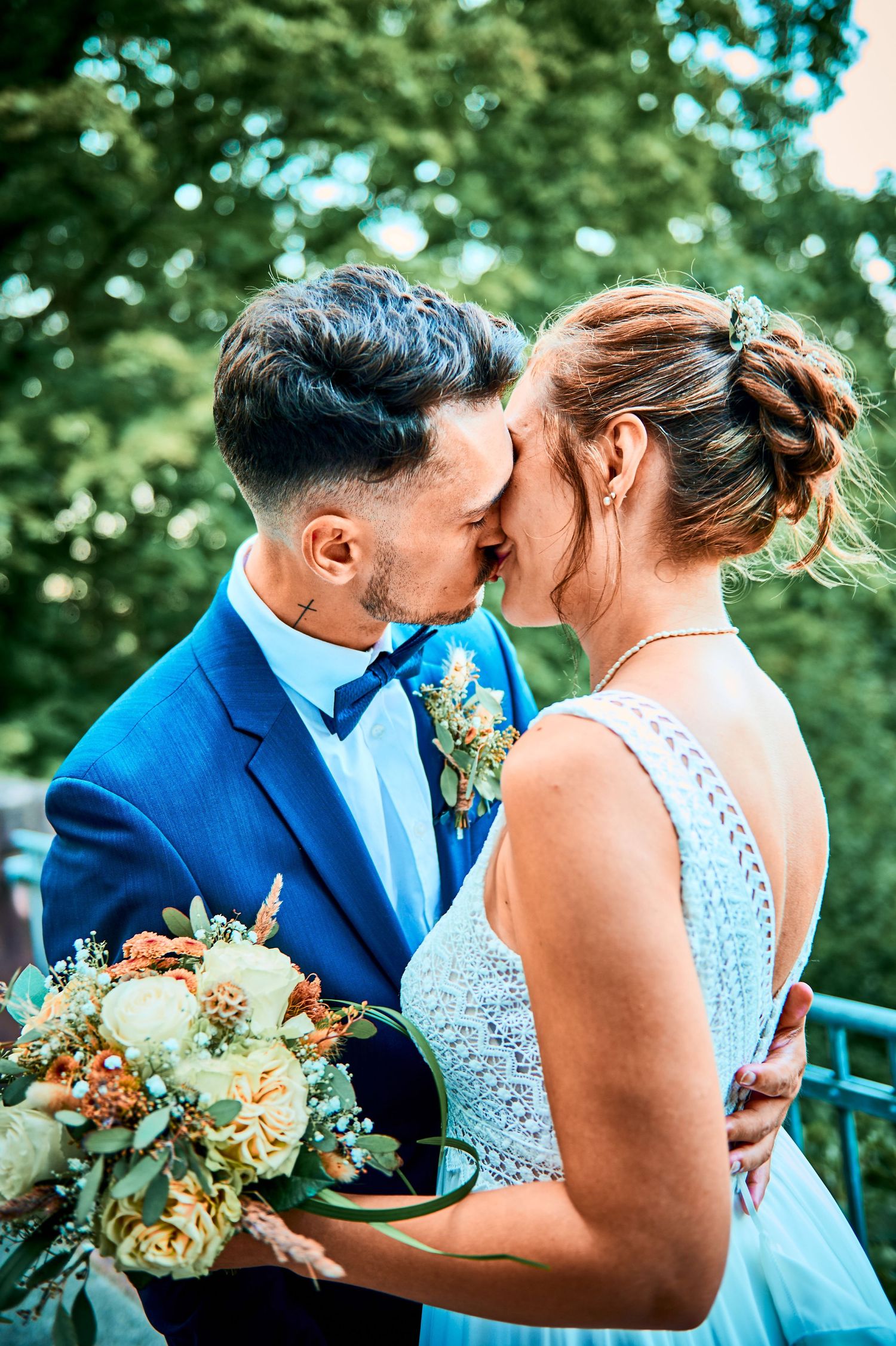 A couple in wedding attire share a romantic kiss outdoors, with the bride holding a peach and cream floral bouquet.