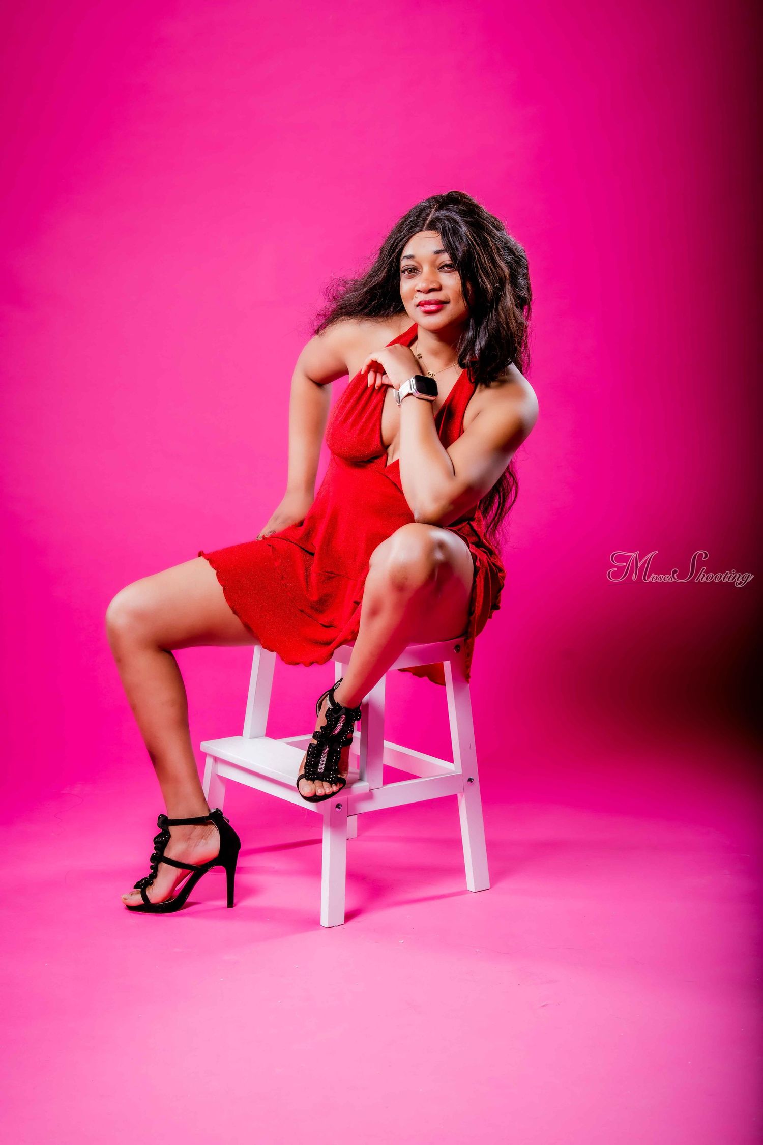 A figure in a red dress poses seductively on a white chair against a vibrant pink studio backdrop.