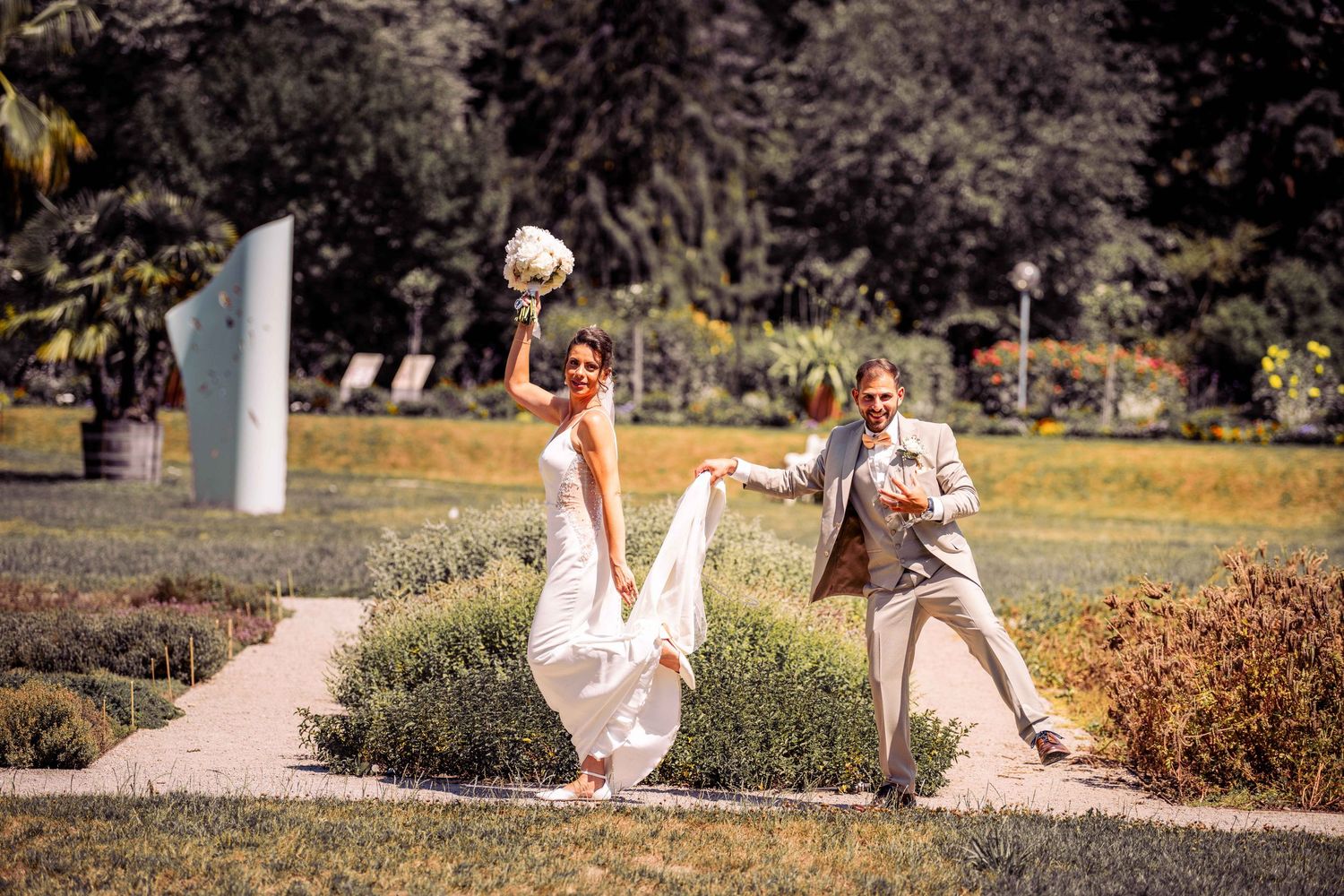 Joyful bride and groom dance playfully in a sunny garden, bride holding bouquet aloft in white dress.
