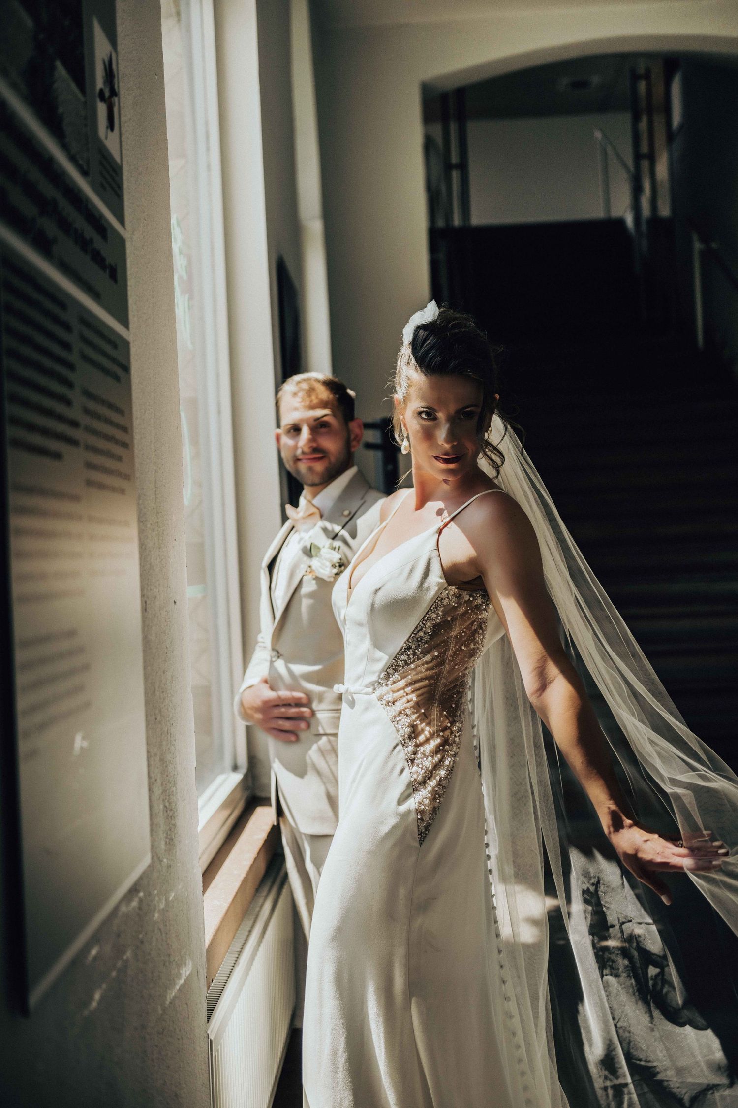 Elegant bride in a white gown with veil stands by a window with groom in a light suit.