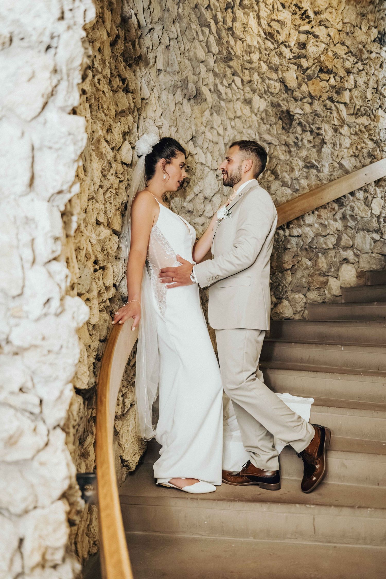 Bride and groom sharing a romantic moment on stone staircase, bride in white gown with veil, groom in beige suit.