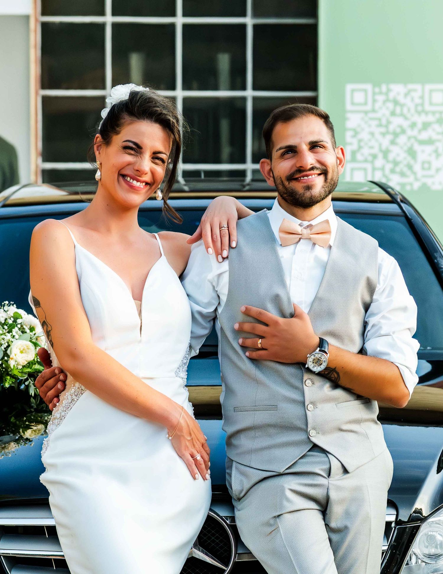 Smiling bride in white gown and groom in gray suit with bow tie pose together leaning on a black car.