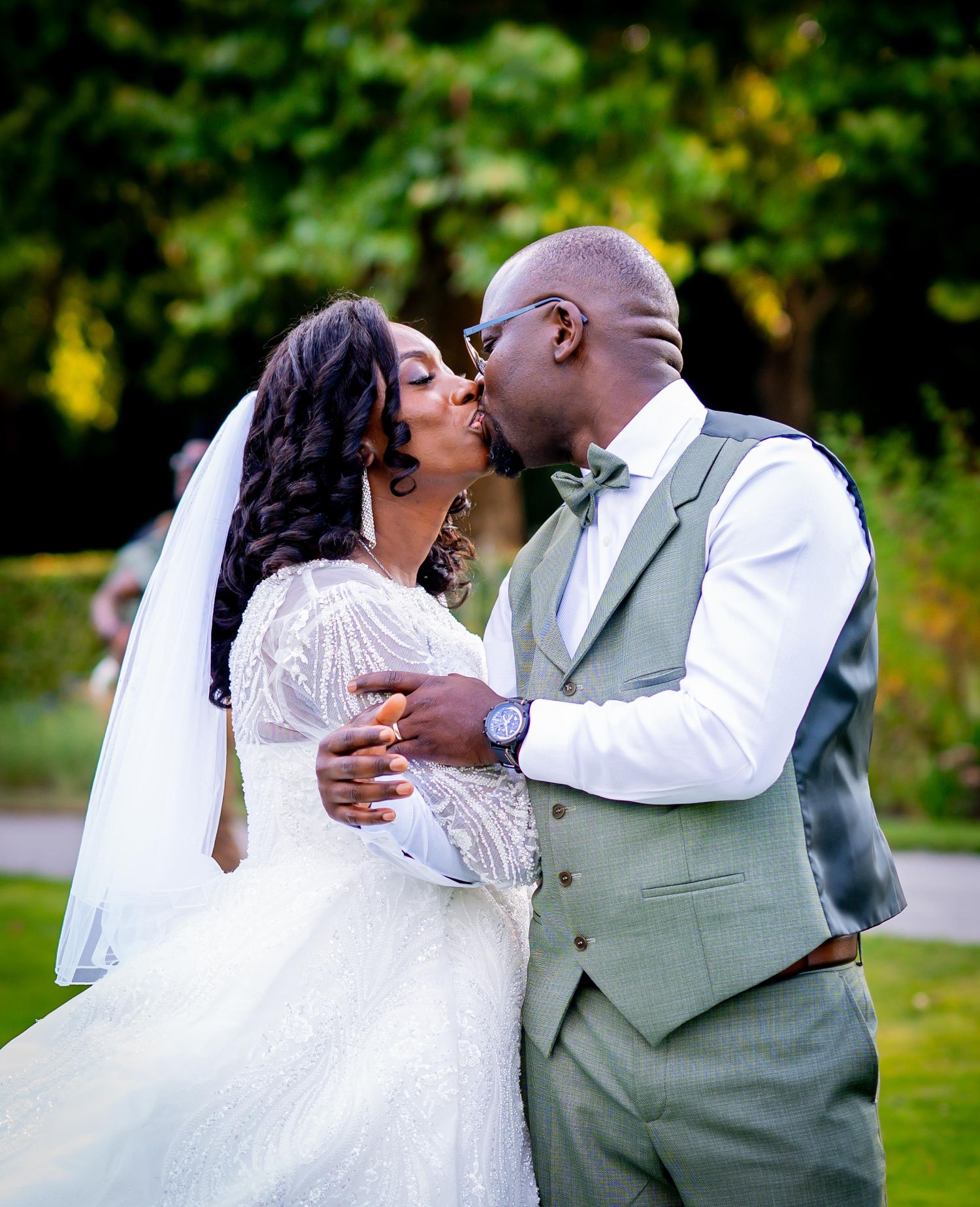 A bride and groom share a romantic kiss outdoors on their wedding day, dressed in a white gown and sage green suit.