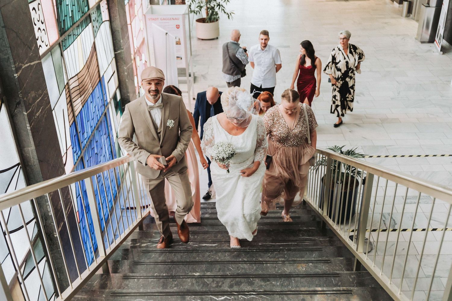Wedding party descends a modern white staircase in a light-filled building interior.