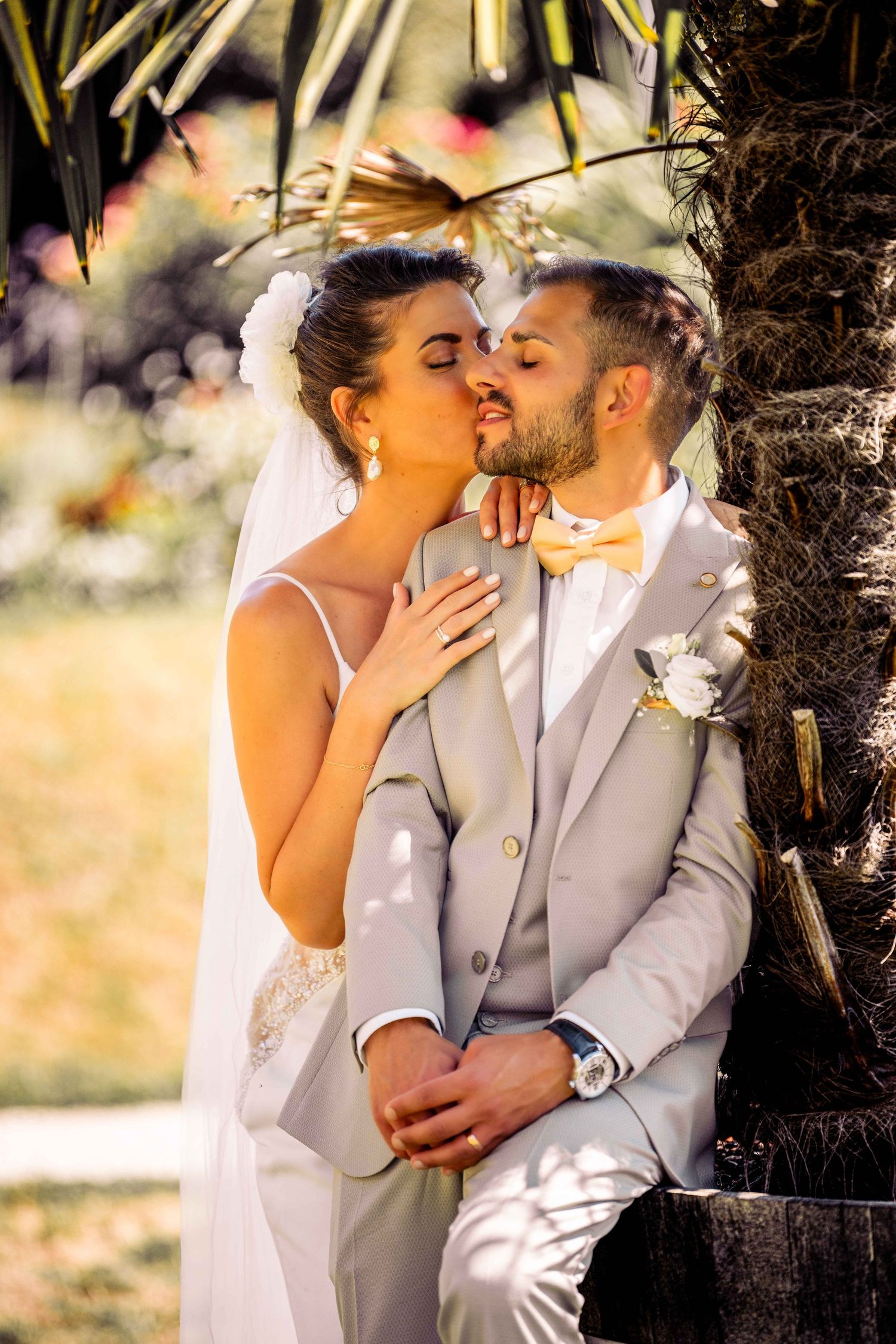 Bride and groom sharing a romantic kiss near a palm tree outdoors on their sunny wedding day.