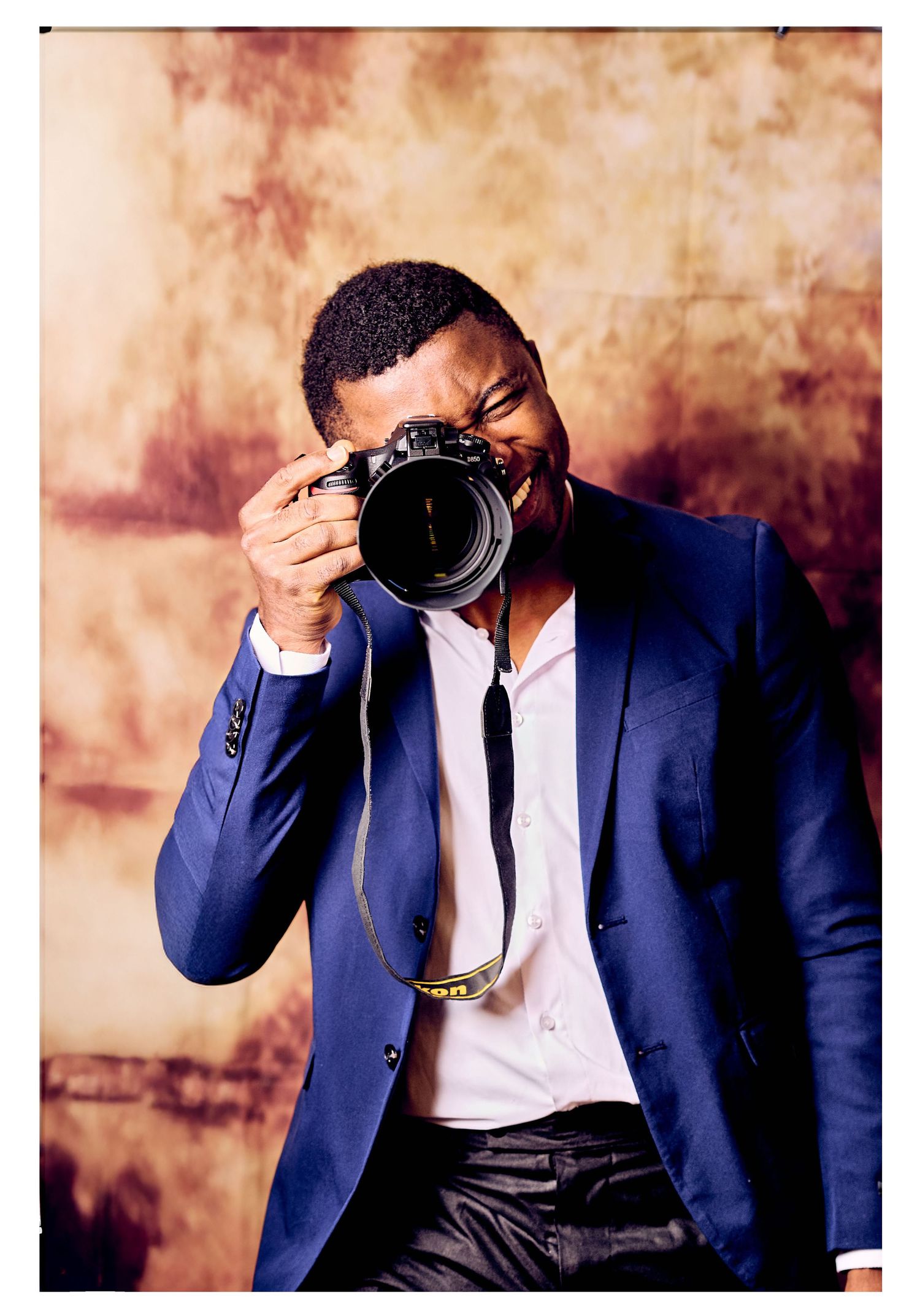 Professional photographer in navy suit and white shirt holds camera while posing against textured brown backdrop.