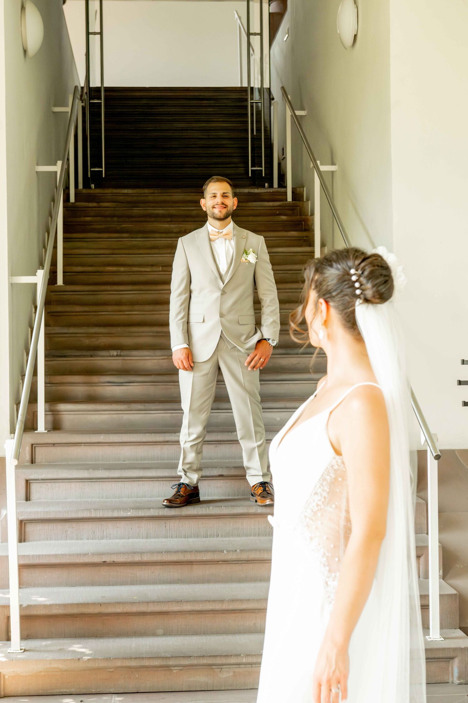 Groom in gray suit smiles at bride in white wedding dress during first look moment on wooden staircase.