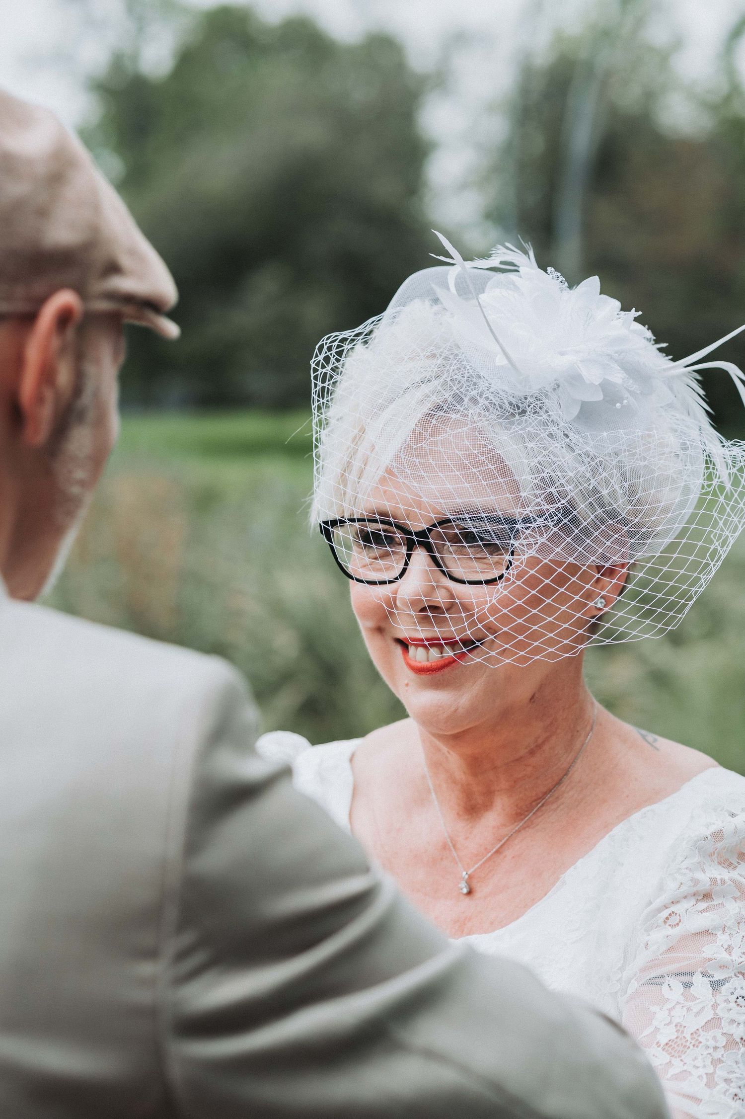 A bride in vintage-inspired lace and birdcage veil smiles joyfully while wearing glasses on her wedding day.