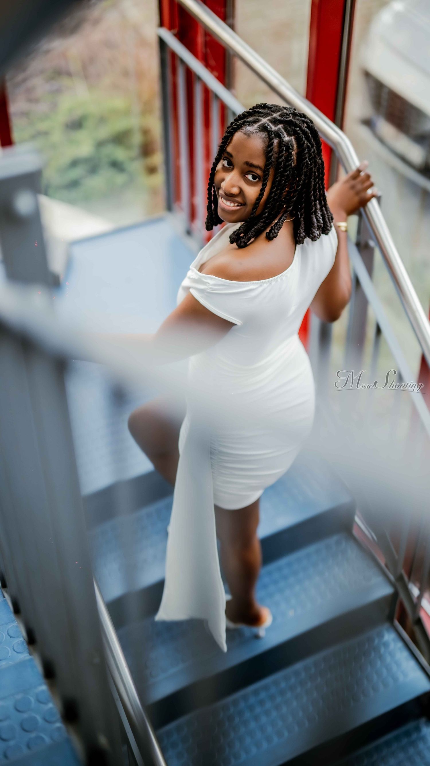 A white form-fitting dress is modeled on industrial metal stairs with red railings.