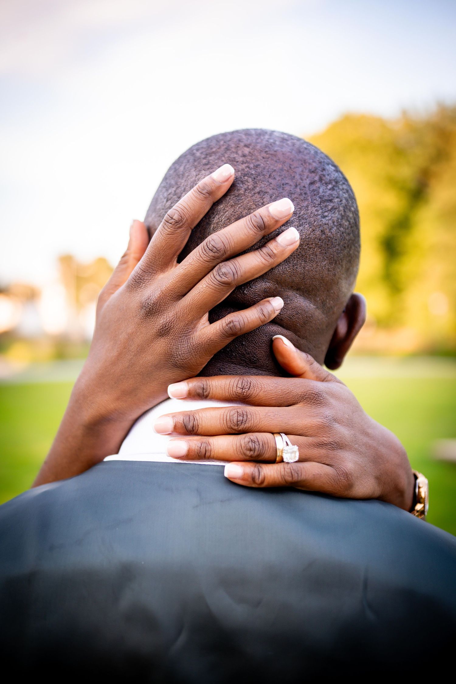 A woman with manicured nails and a diamond ring tenderly cradles a mans head from behind in an outdoor setting.