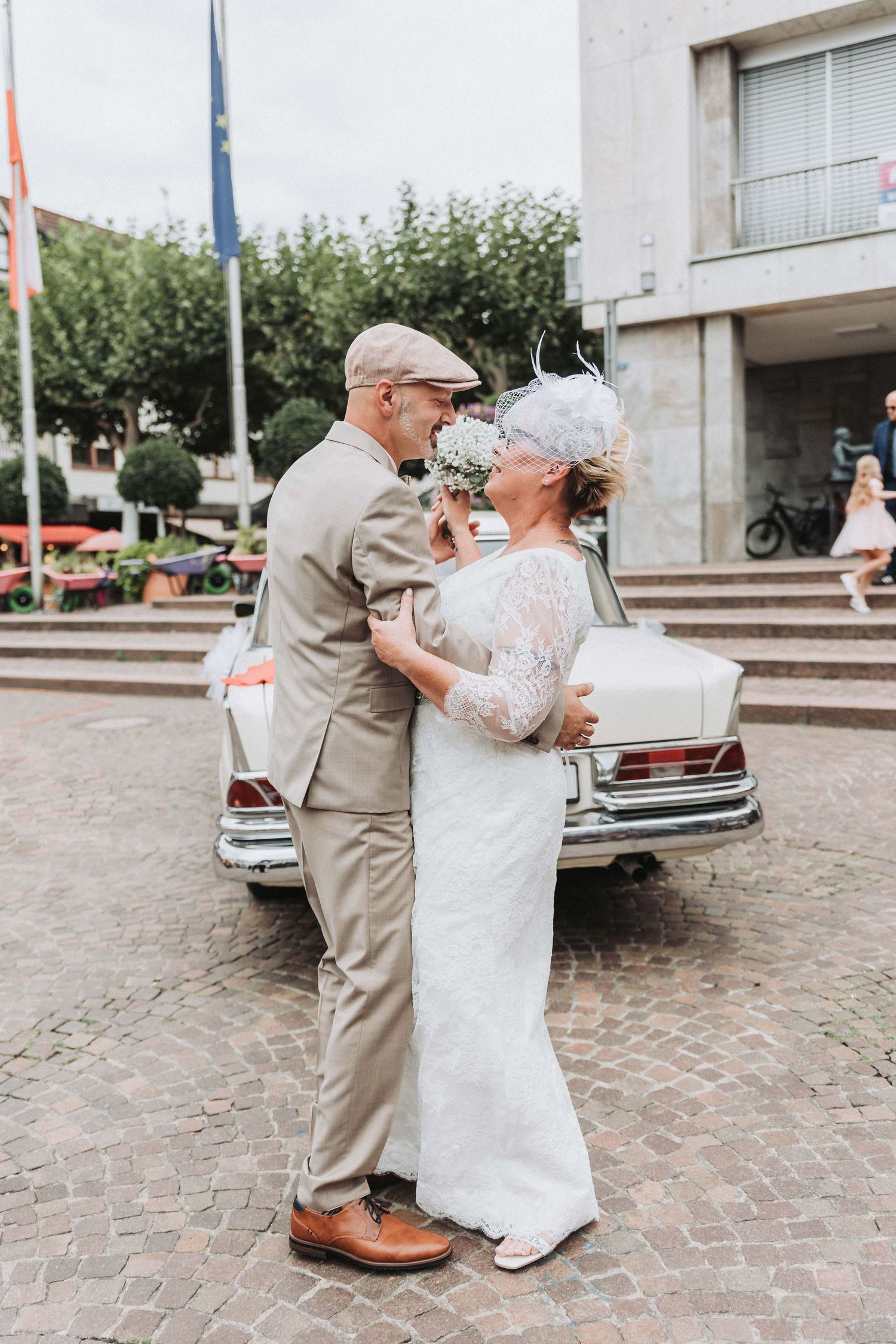 A couple in wedding attire dancing together on a cobblestone courtyard with vintage cars in the background.