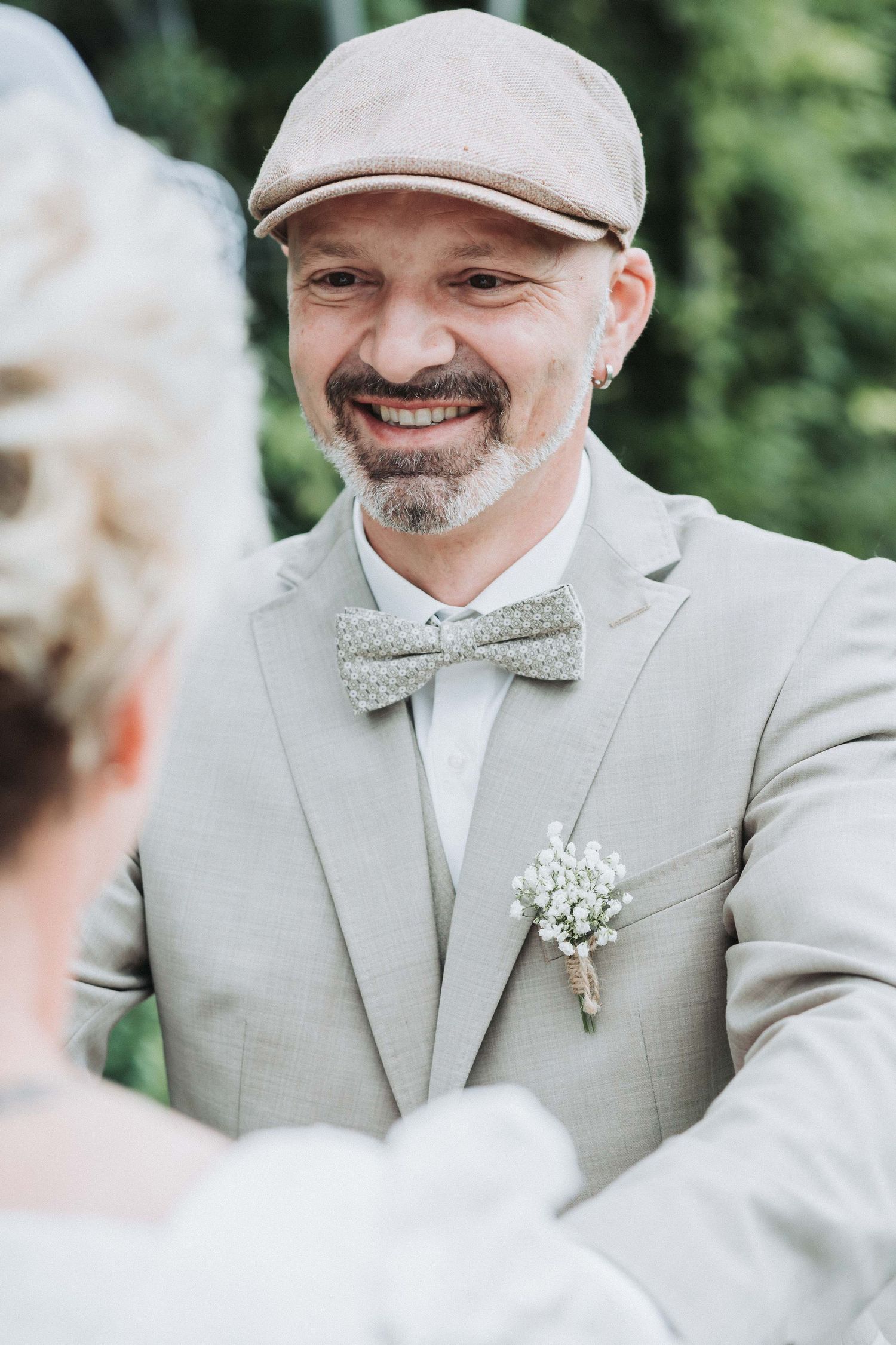 A groom in a light gray suit and polka dot bowtie shares a joyful moment during an outdoor wedding ceremony.
