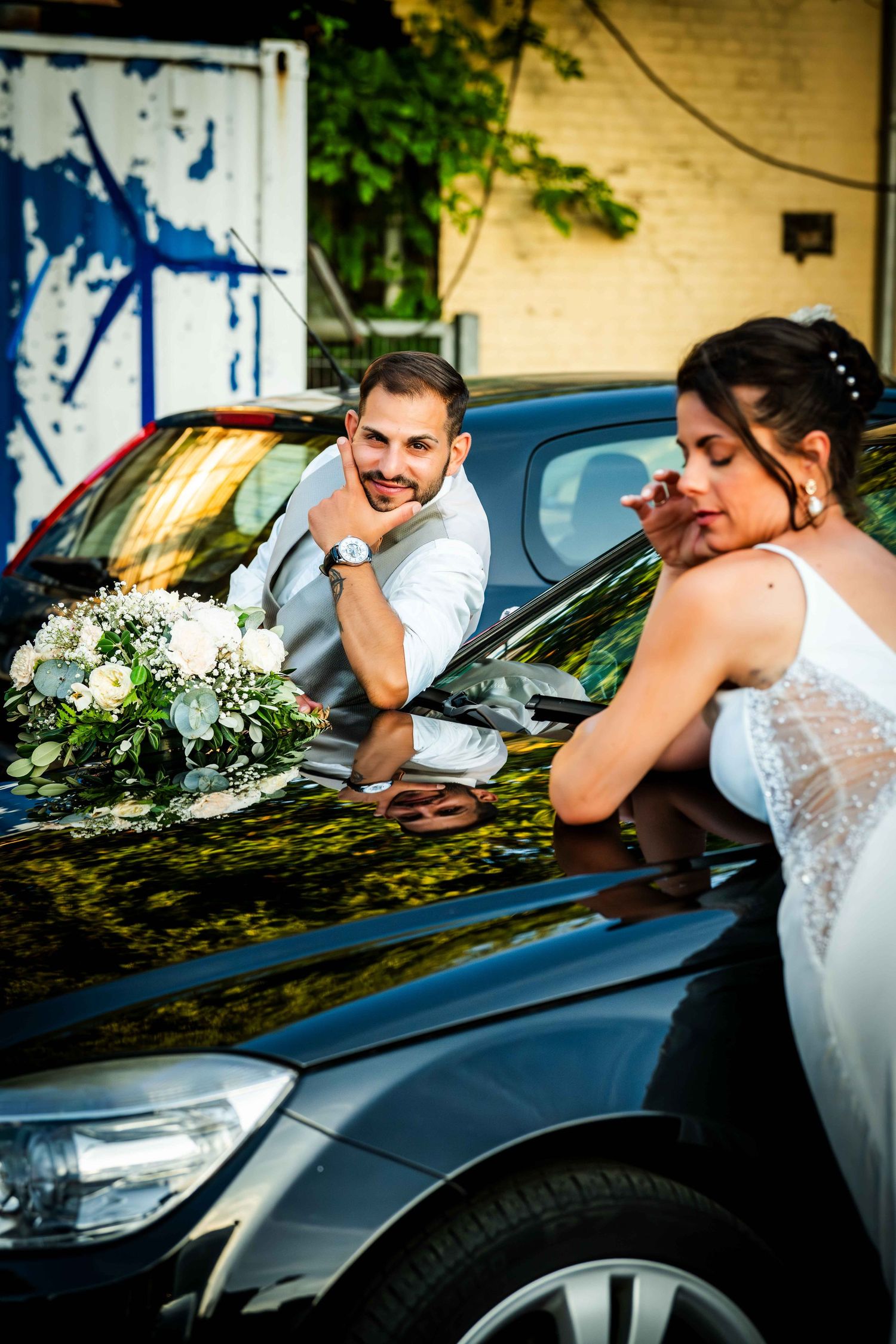 Bride and groom leaning on black car hood with white flower bouquet, sharing a romantic moment outdoors.