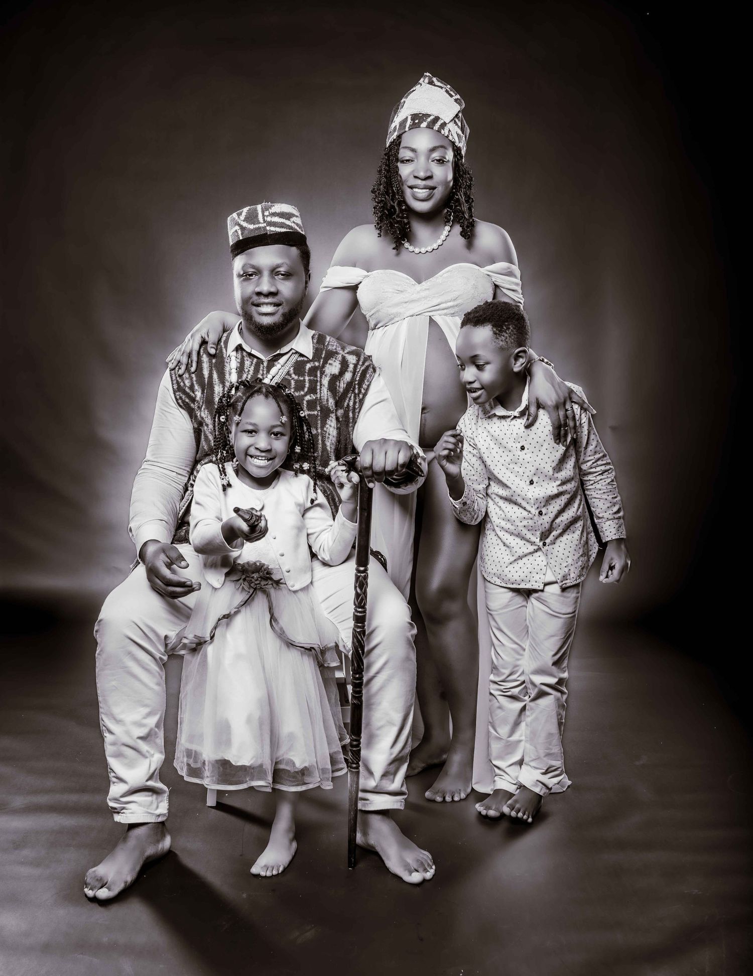 A black and white studio portrait of a family of four wearing coordinated light-colored outfits against a dark background.