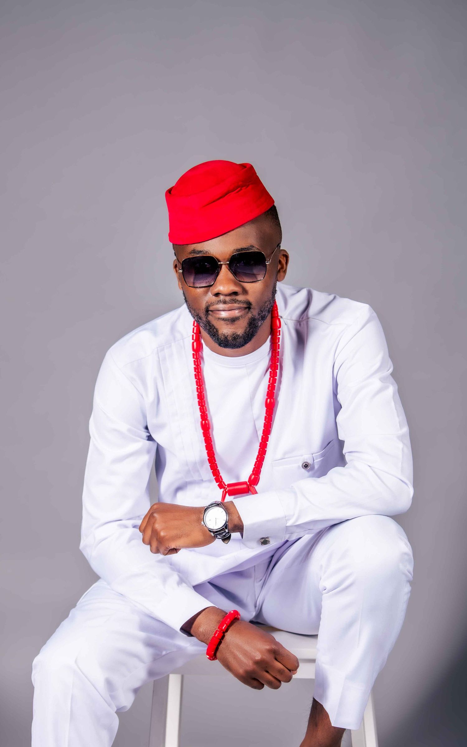A person in all-white traditional attire with red beads and cap poses for a professional photo series.