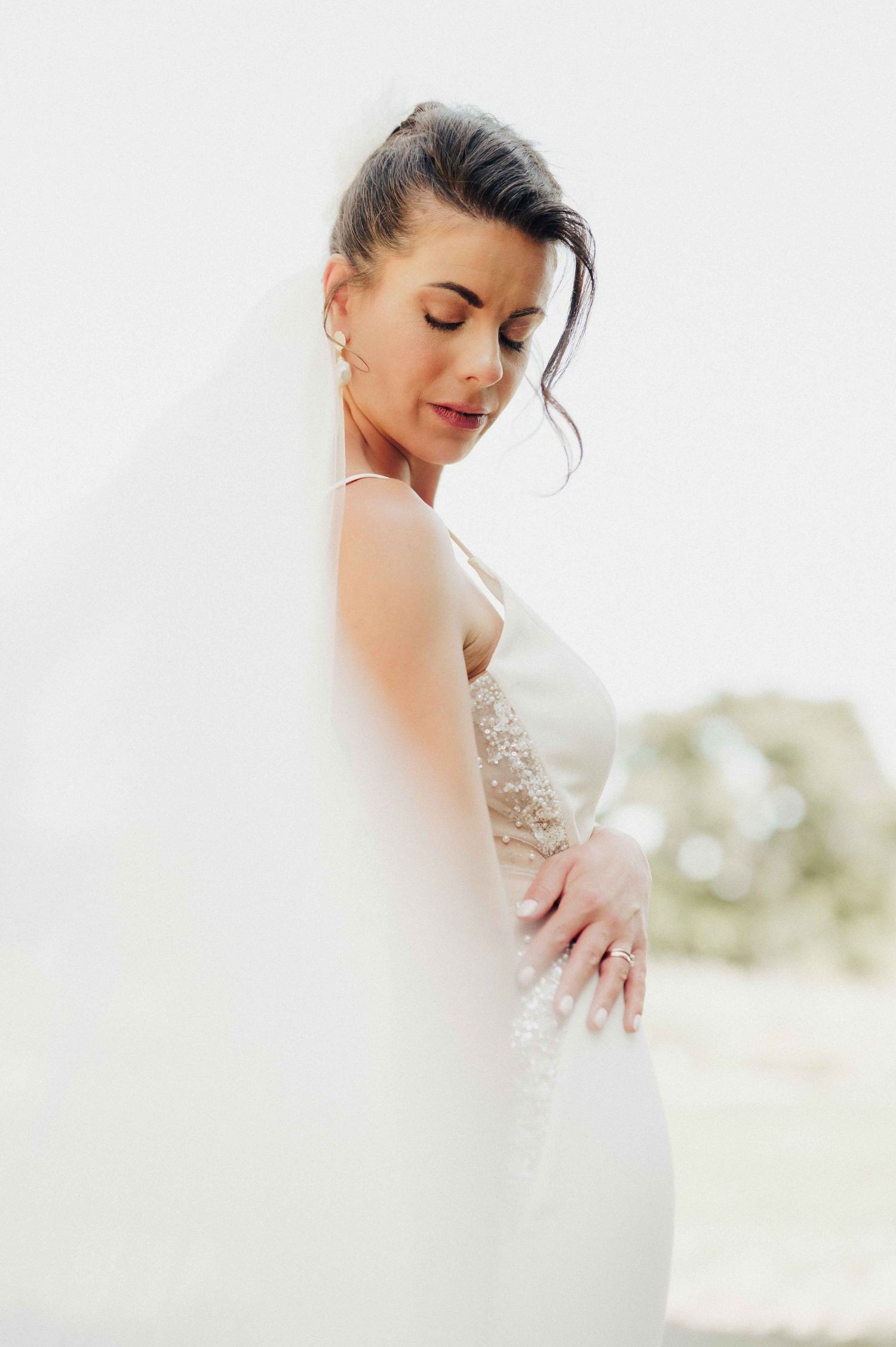 Elegant bride in a white lace gown and veil, eyes closed, posing gracefully against a soft white background.
