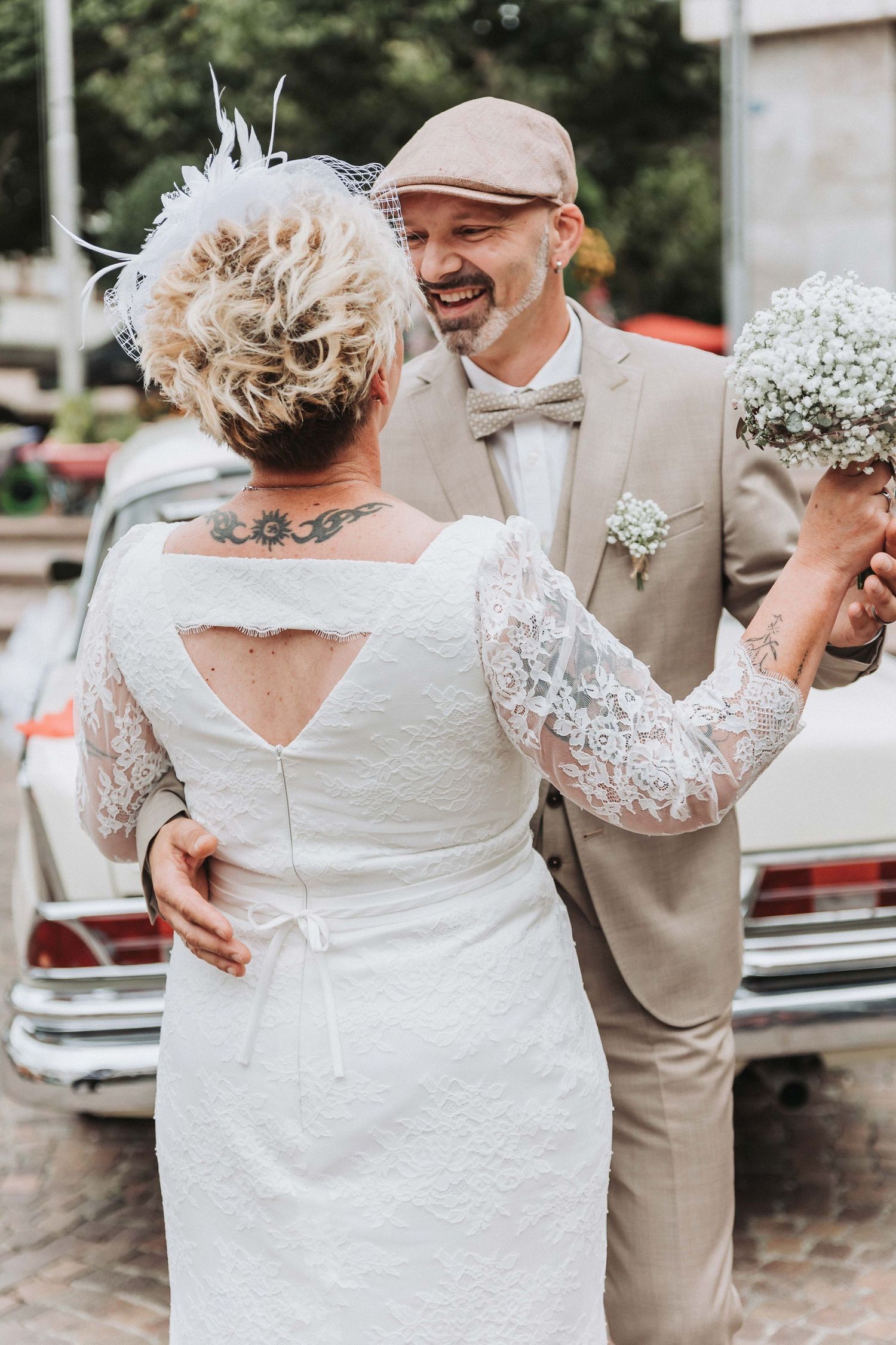 A wedding couple shares a joyful dance moment outdoors, with the bride wearing a lace-sleeved white dress.