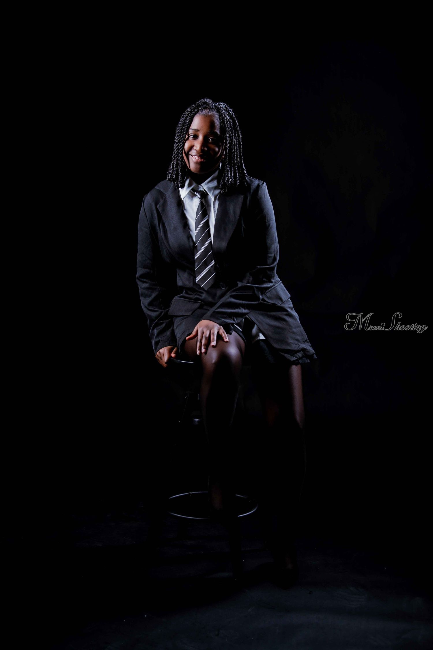 Professional studio portrait of someone in a dark business suit and tie sitting on a stool against a black background.
