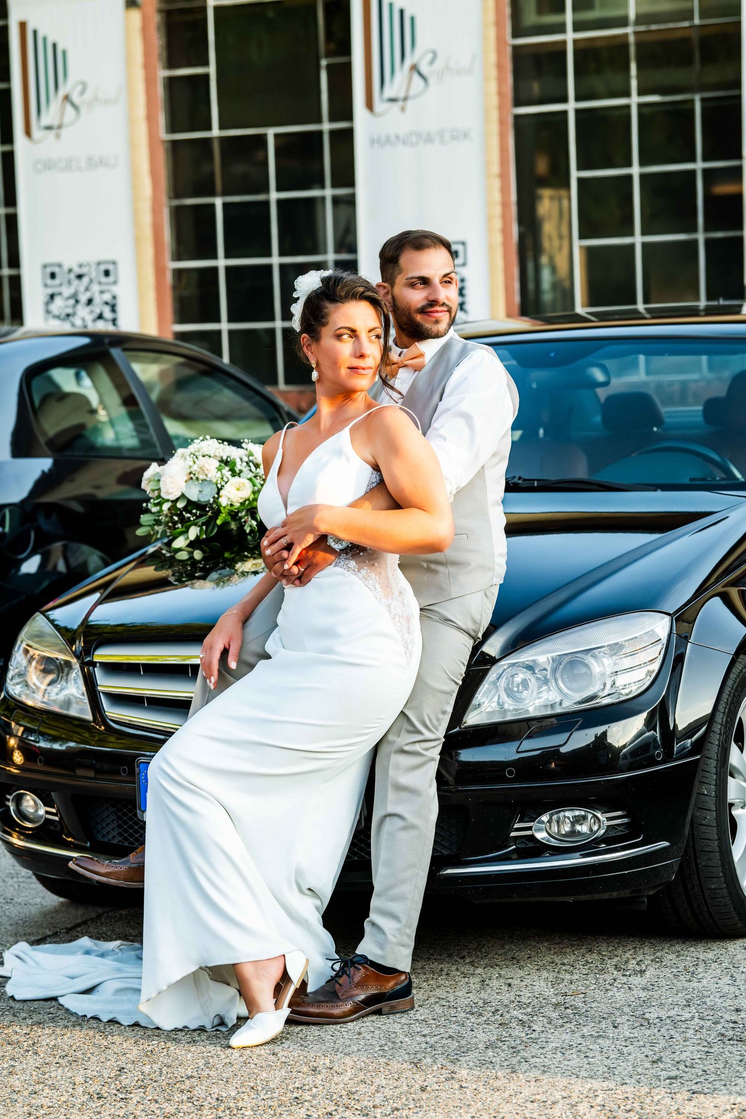 Wedding couple posing by a black Mercedes car, bride in white gown holding bouquet, groom in gray vest suit.
