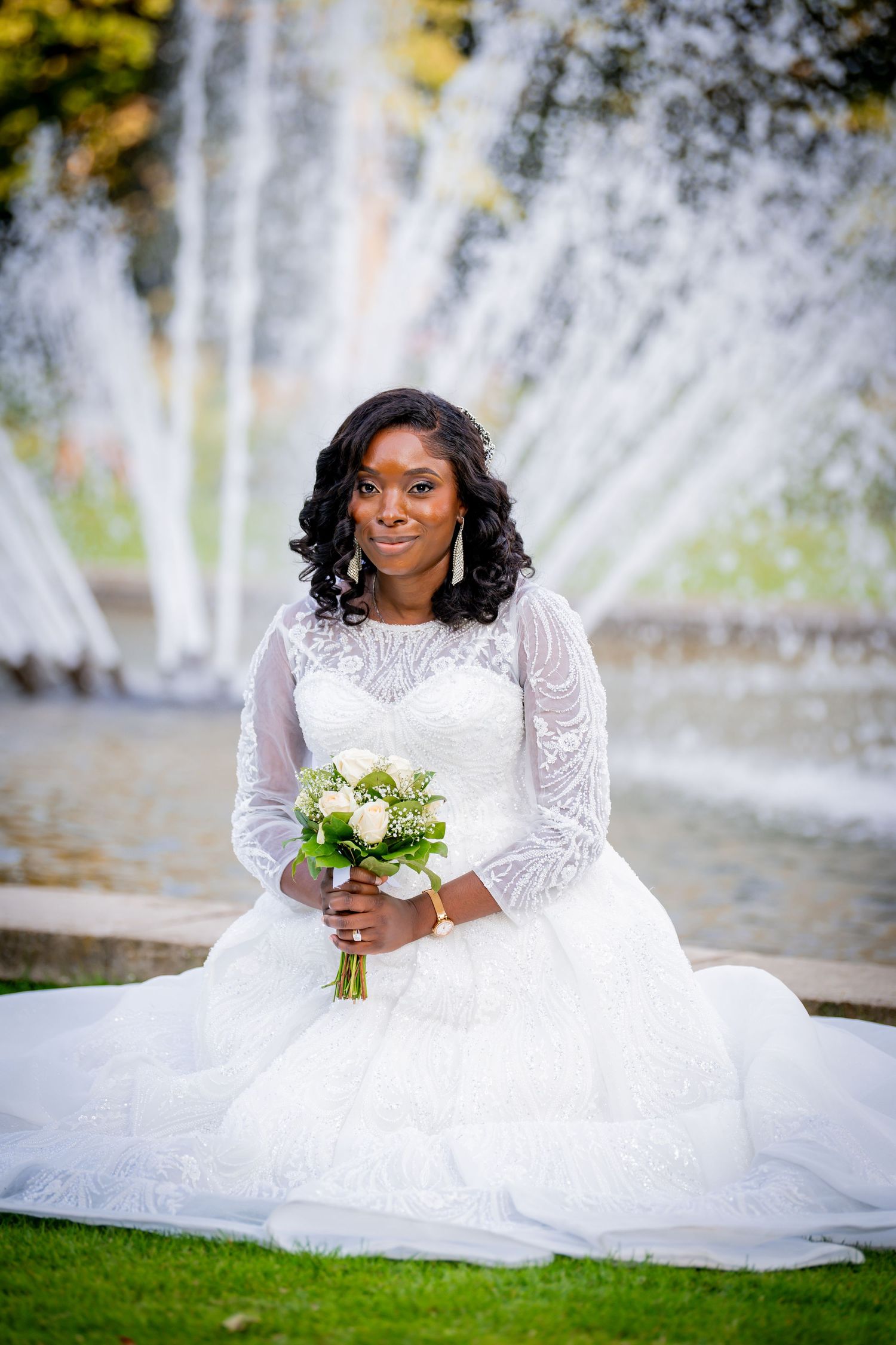 Bride in white lace long-sleeve gown sits on grass holding bouquet before fountain.