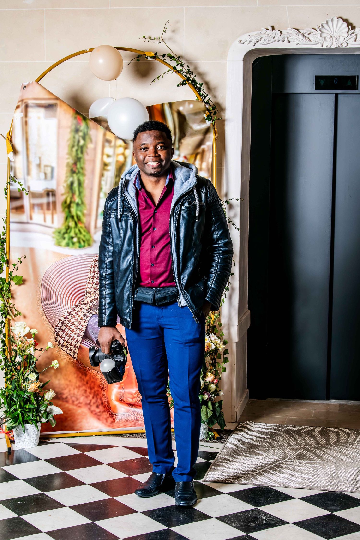 A person in a leather jacket and blue pants poses on a black and white checkered floor with holiday decorations.