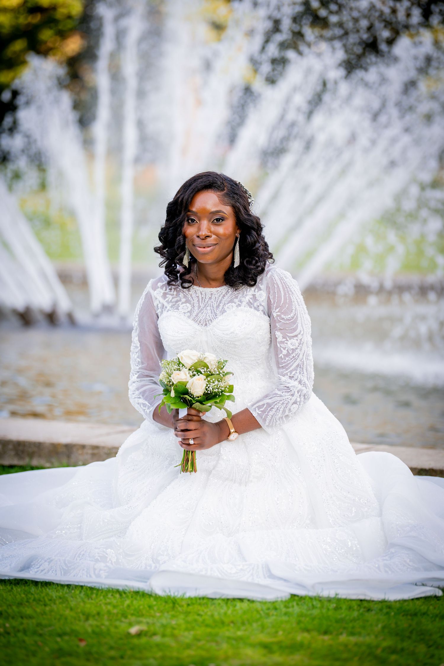 Smiling bride in white lace gown holds bouquet, seated on grass before a fountain.
