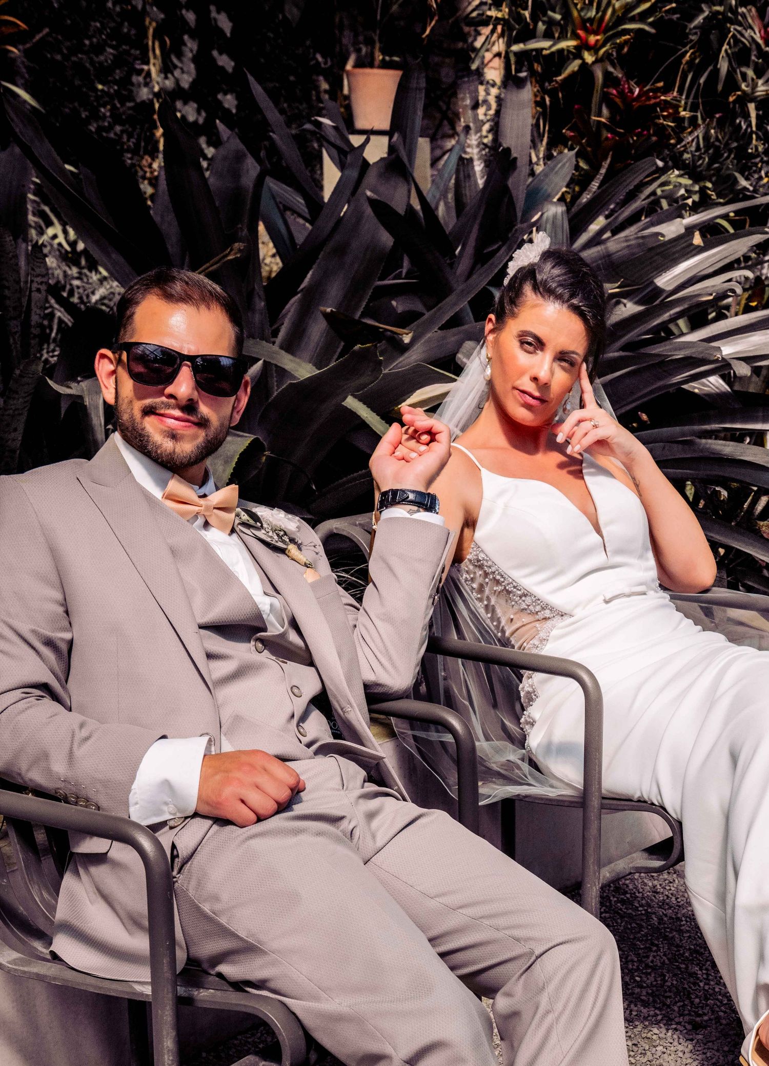 Bride and groom in elegant wedding attire relaxing in chairs outdoors, groom in gray suit with bow tie and sunglasses.