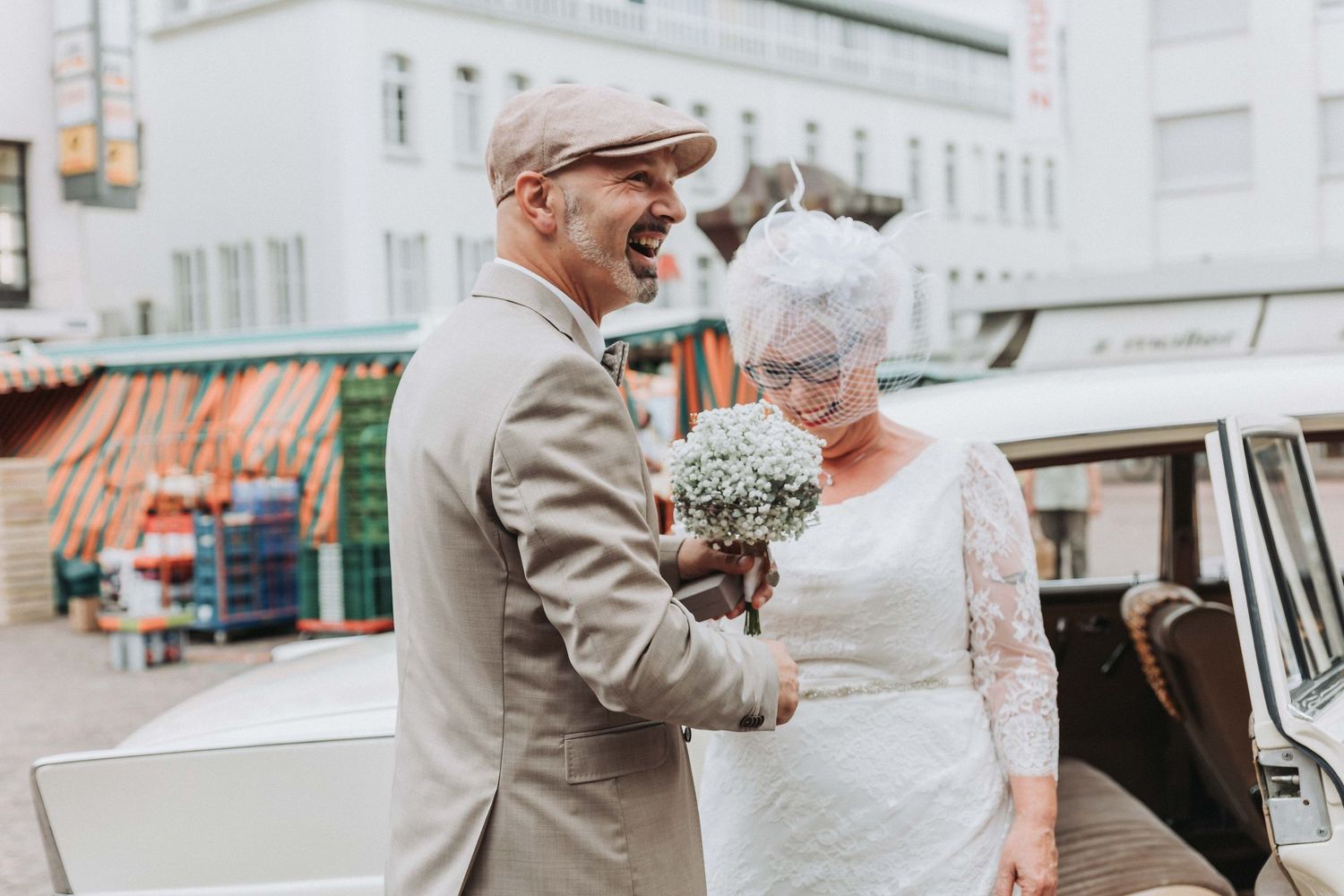 A vintage-inspired wedding couple embraces on a city street, wearing a flat cap and lace dress with classic styling.