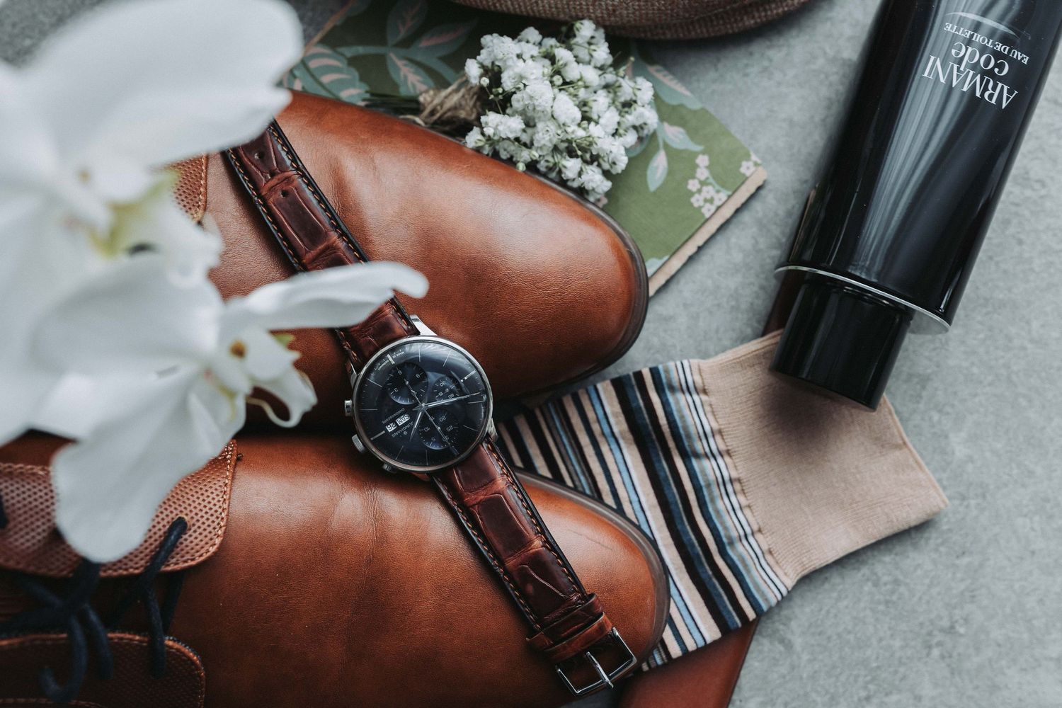 Elegant wedding details with brown leather shoes, white orchids, and accessories arranged on a gray surface.