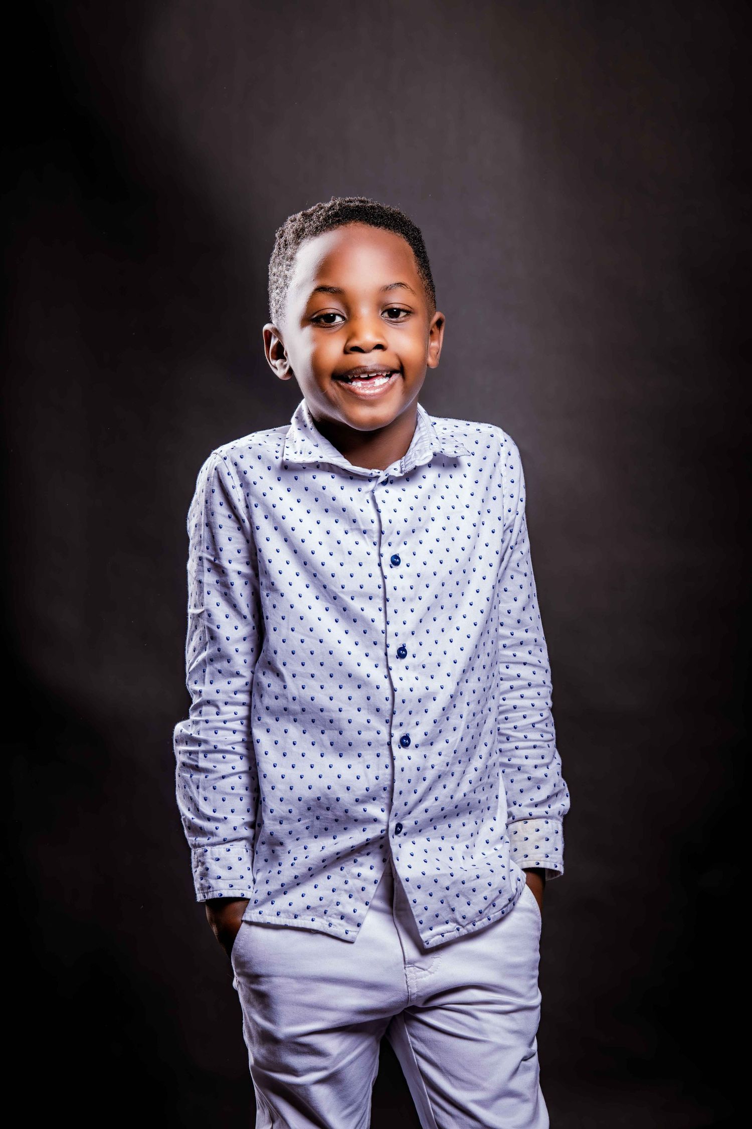 A child wearing a polka dot dress shirt and light gray pants poses against a dark background in a portrait series.