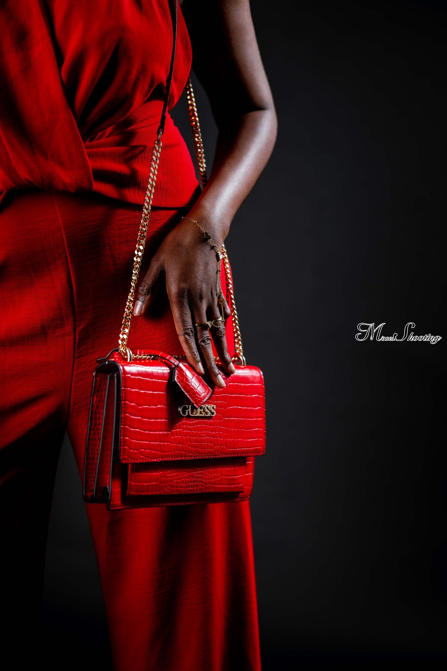 Elegant red crocodile-textured handbag with gold chain strap held against flowing red dress on dark background.