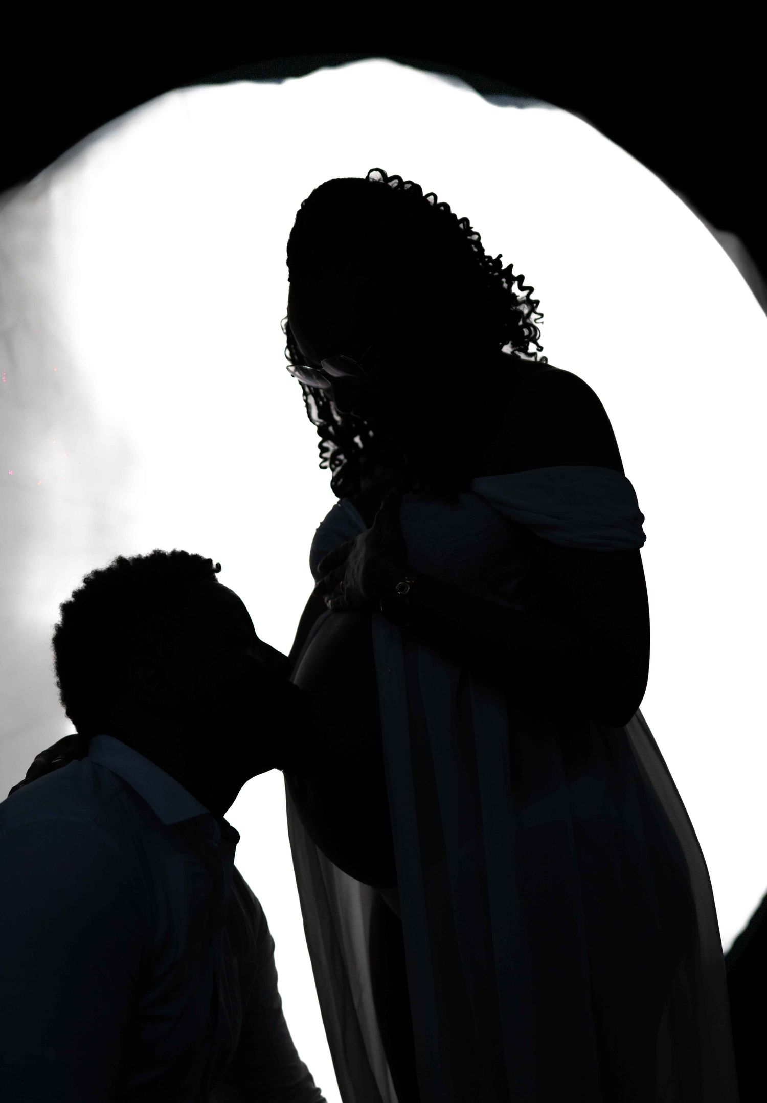 Silhouetted figures pose dramatically against a bright circular light creating a romantic maternity portrait.