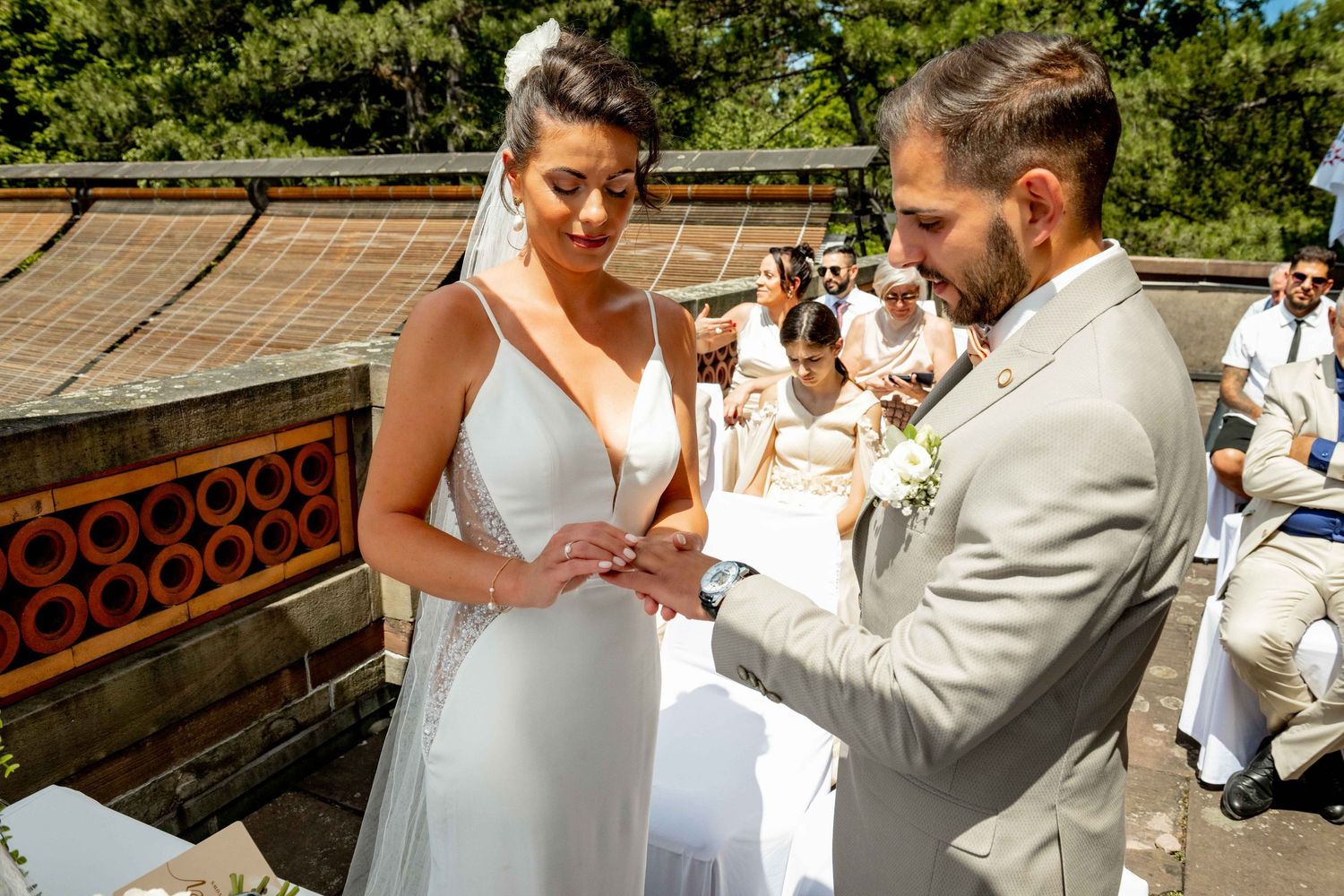 Bride and groom exchanging rings during outdoor wedding ceremony with guests seated in background.