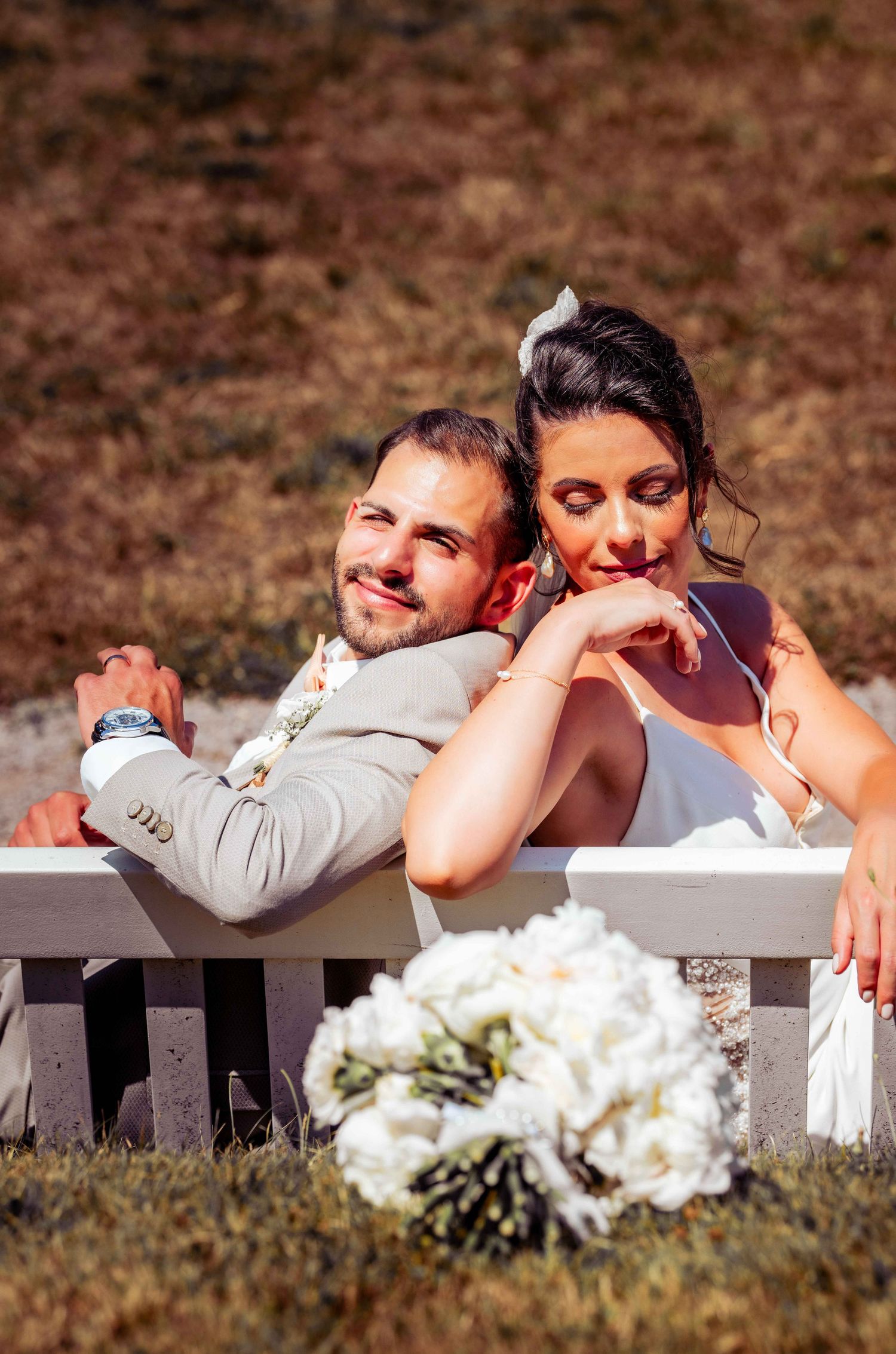 Romantic bride leaning on groom's shoulder on white bench with white wedding bouquet in foreground.
