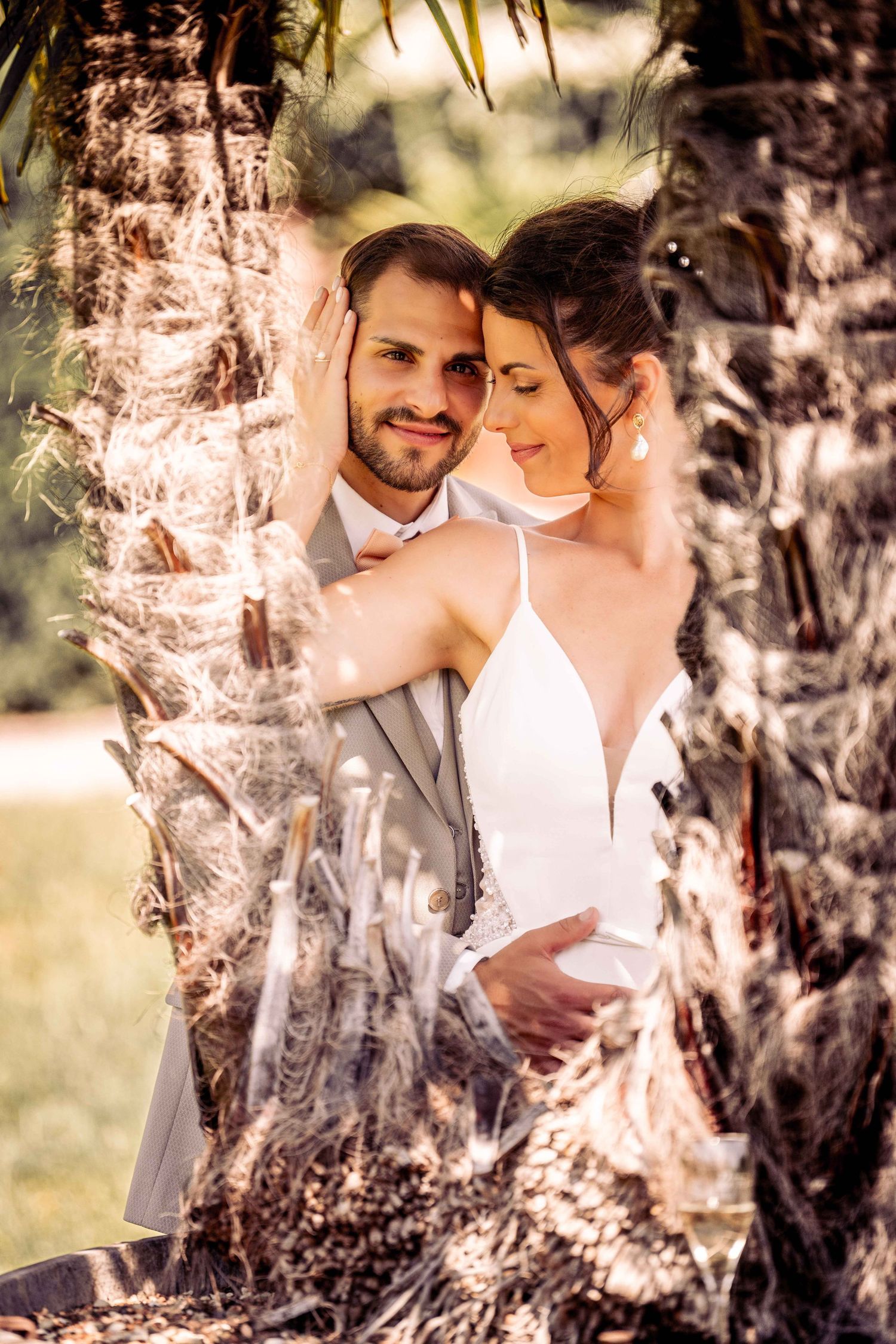 Wedding couple embracing tenderly between palm trees in warm sunlight, bride in white halter gown.