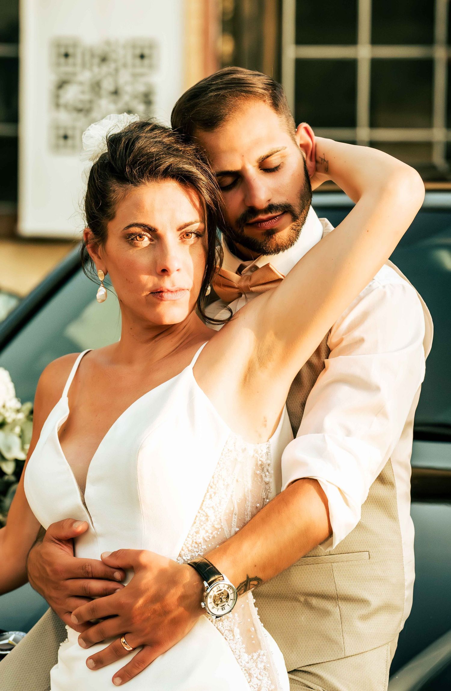 A bride in a white gown and groom in a tan suit embrace tenderly in a romantic outdoor wedding portrait.