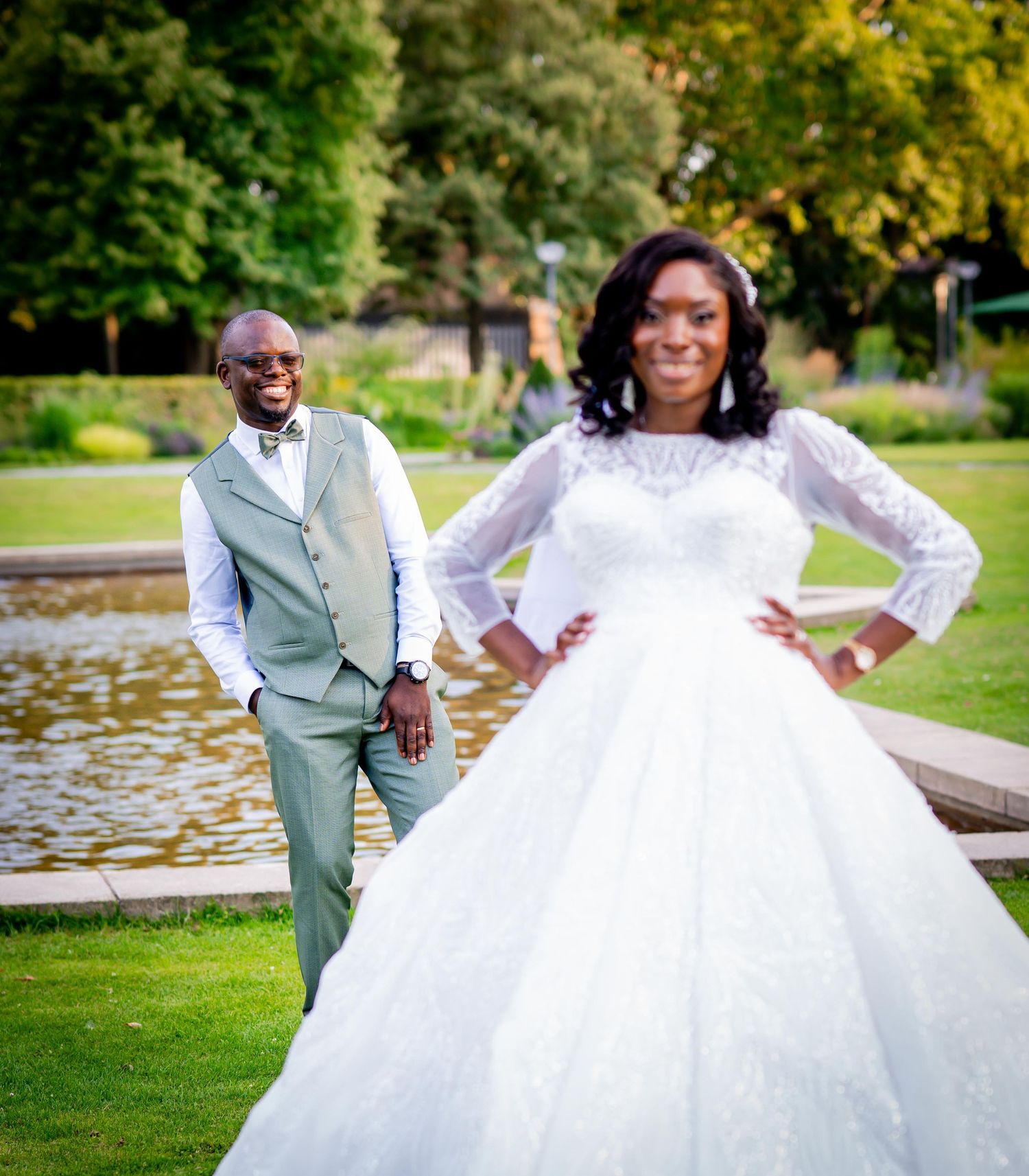 Black bride in white lace gown poses confidently with groom by pond in park.